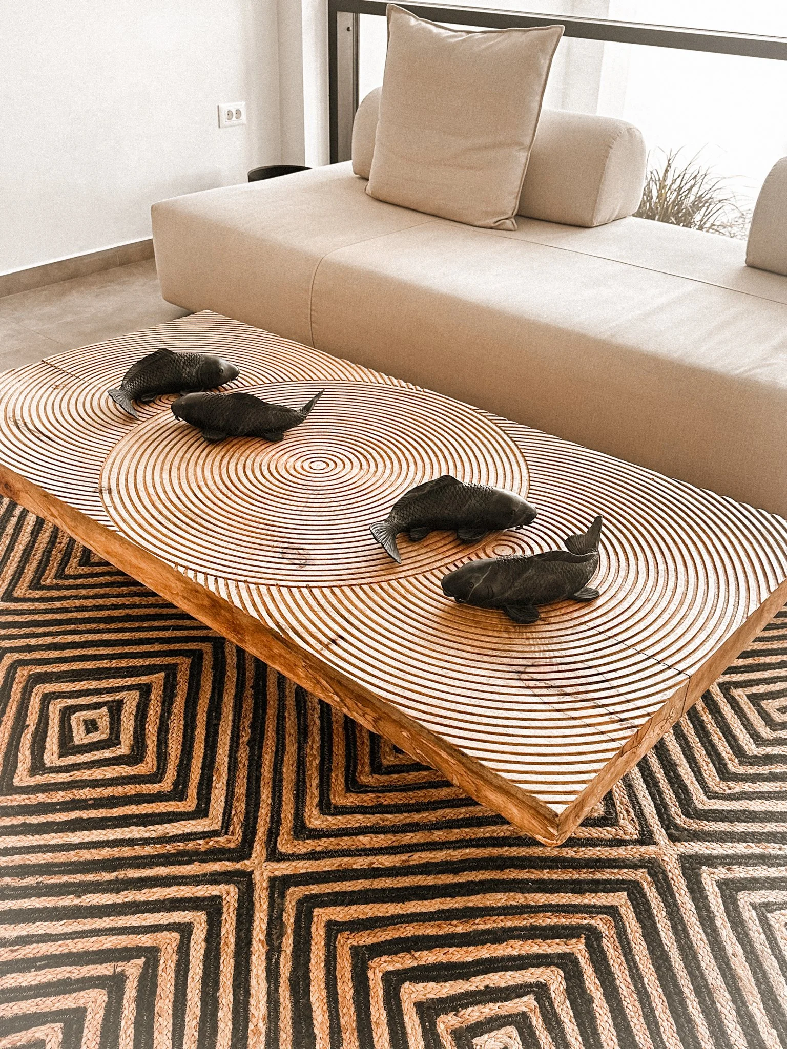Living room with beige sofa, decorative pillows, charred wood coffee table with carved concentric circles, black fish sculptures, and patterned area rug.