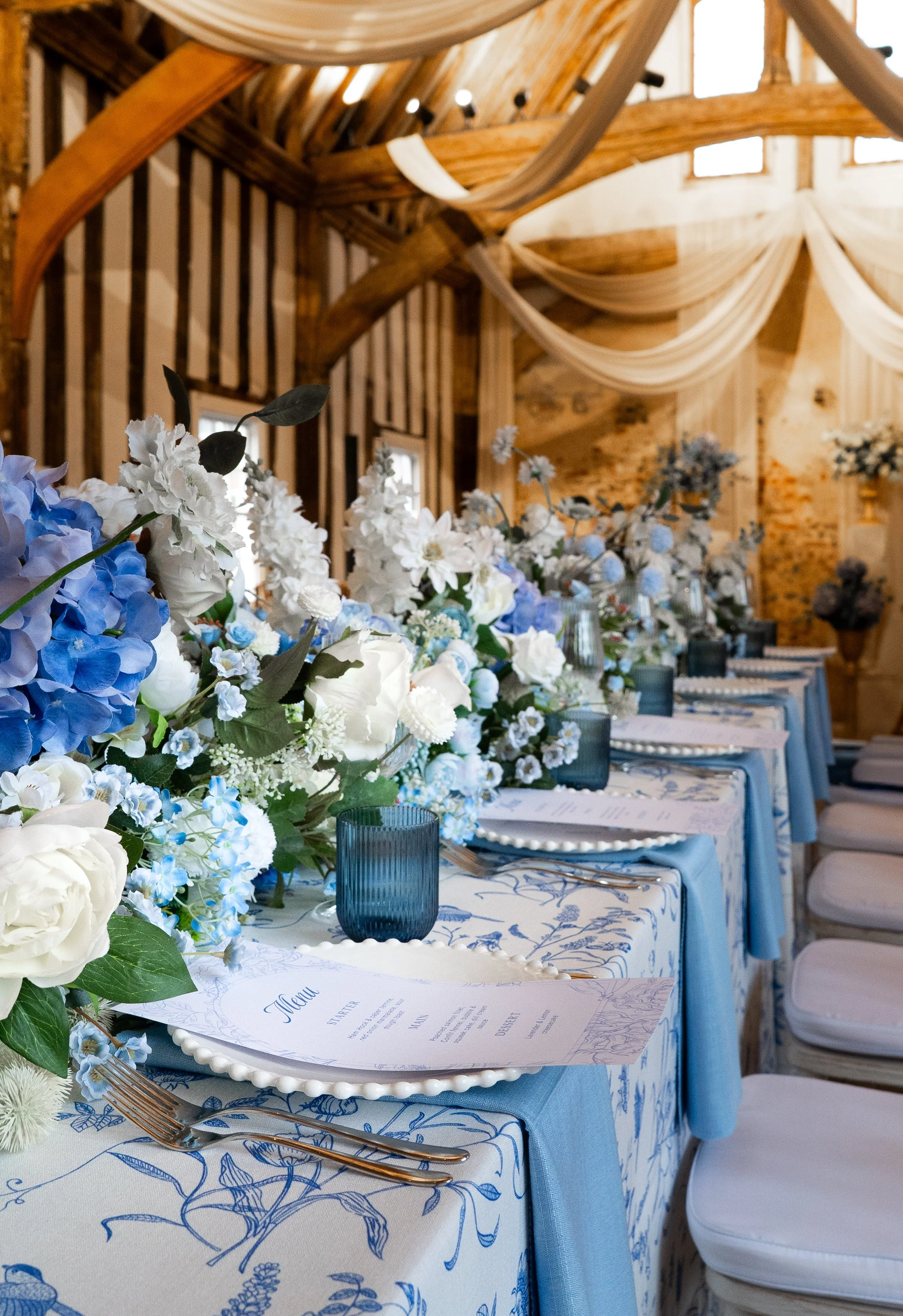 A decorated banquet table with floral centerpieces, menus, and blue glasses set in a rustic wooden venue with draped fabric and natural wood beams.