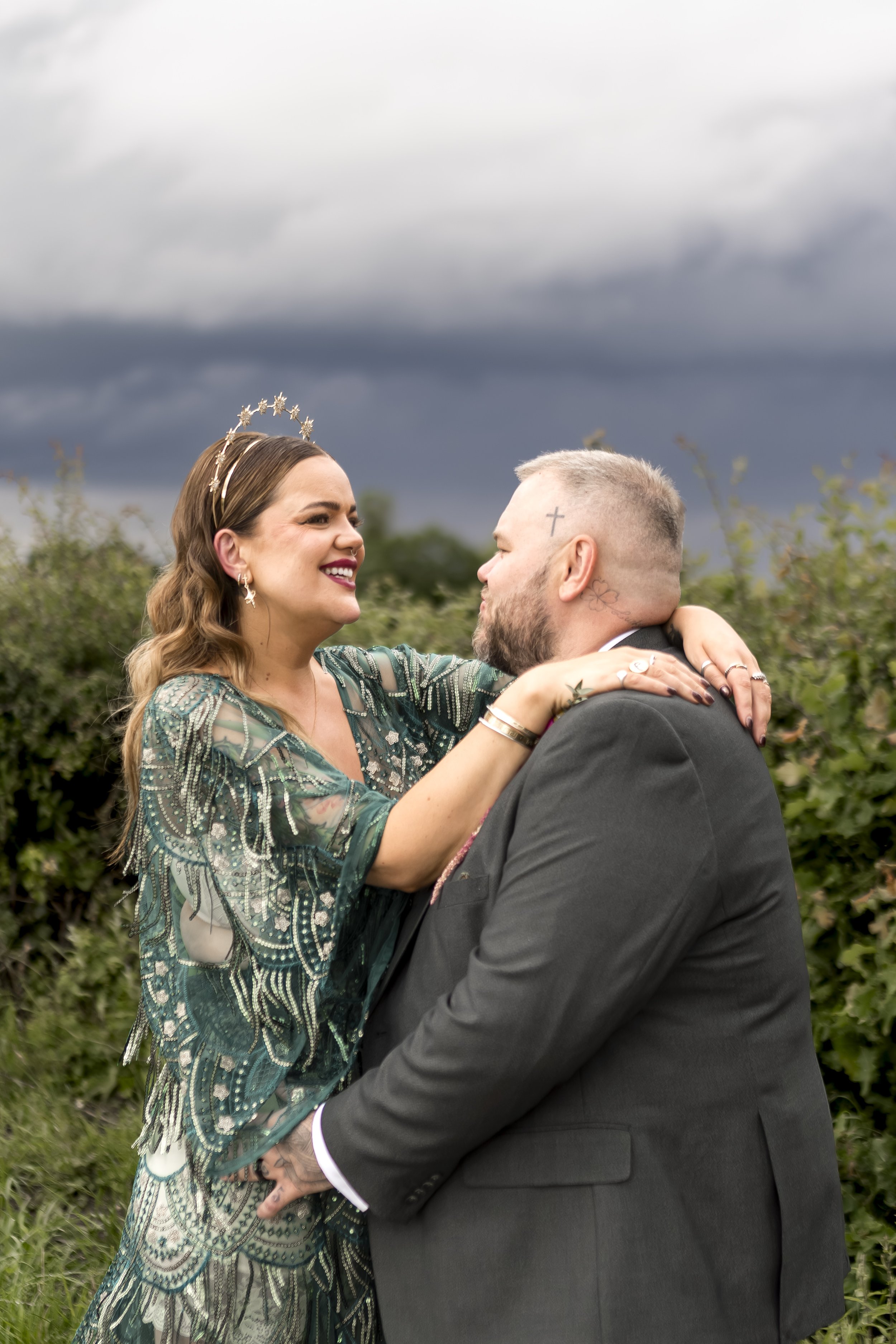 A couple is dancing outdoors under a cloudy sky. The woman is wearing a green dress with intricate patterns, a tiara, and jewelry, and is smiling at the man. The man has a beard, short hair, a cross tattoo on his head, and is wearing a gray suit. The