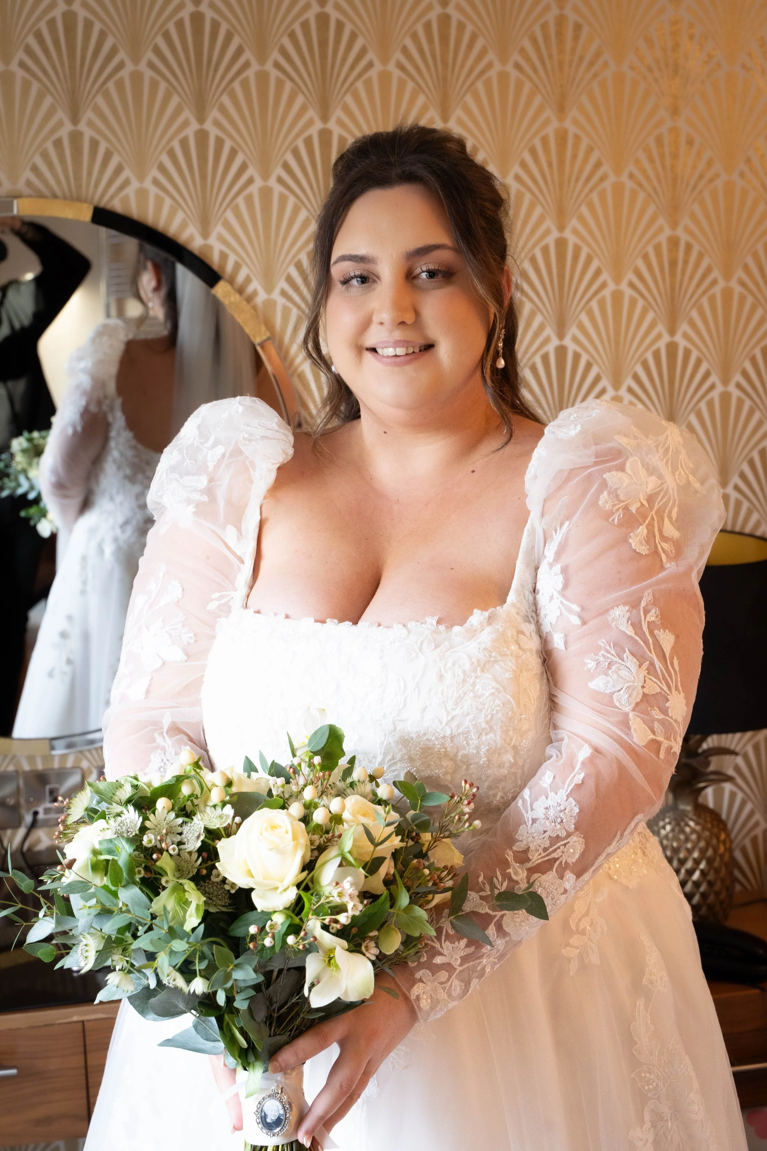 A woman in a white wedding dress holds a bouquet of white and green flowers, standing in front of a decorative golden art deco wallpaper, with her reflection visible in a mirror behind her.