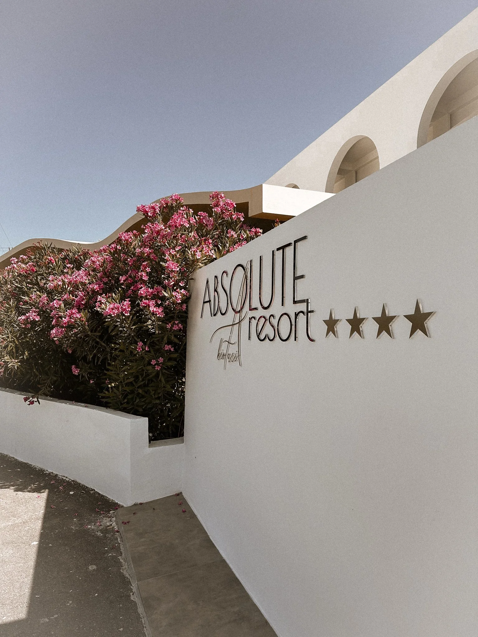 White sign with black text reading "ABSOLUTE resort" and three stars underneath, with pink flowering shrubbery and a modern building with arches in the background under a clear sky.