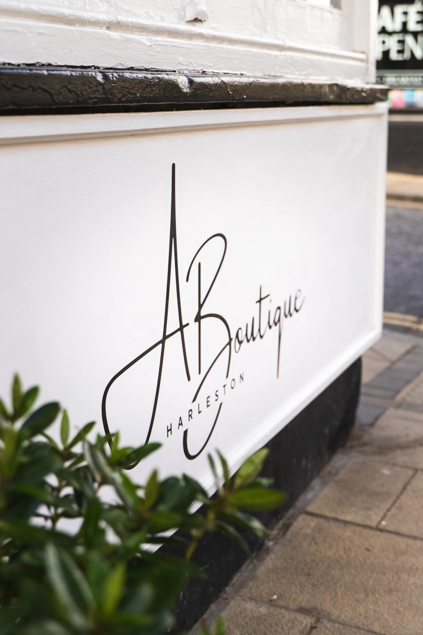 Close-up of a white sign with black cursive writing that says "A Boutique Charleston" on a sidewalk, with greenery in the foreground and a blurred storefront in the background.