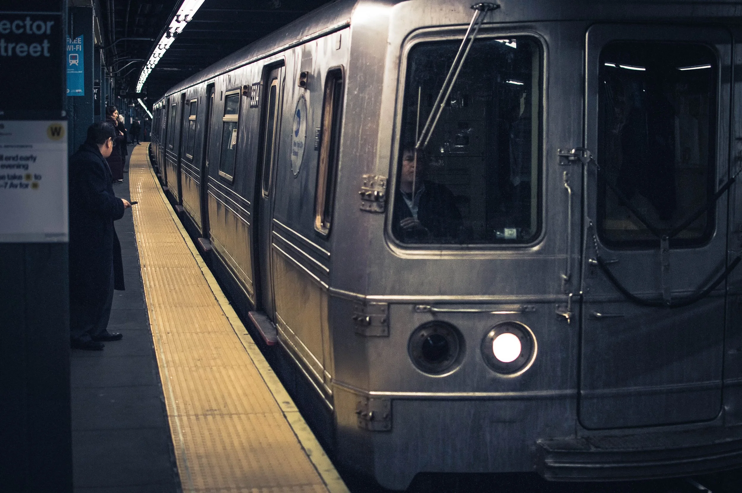 A subway train at a station platform with a man looking at his phone in the foreground and other passengers in the background, under a curved ceiling with bright overhead lights.