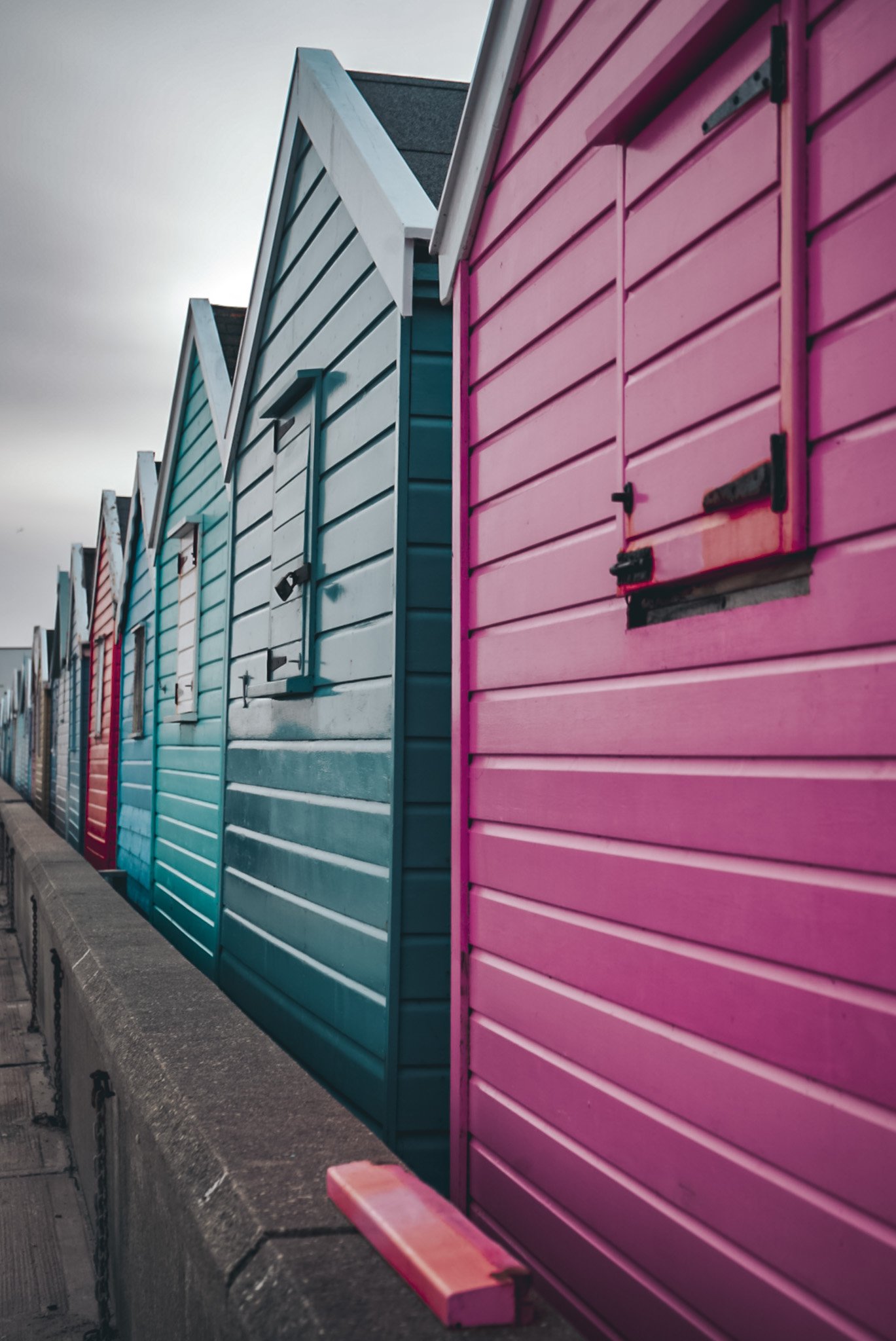 Colorful beach huts with pink, blue, and red panels, situated along a concrete walkway.
