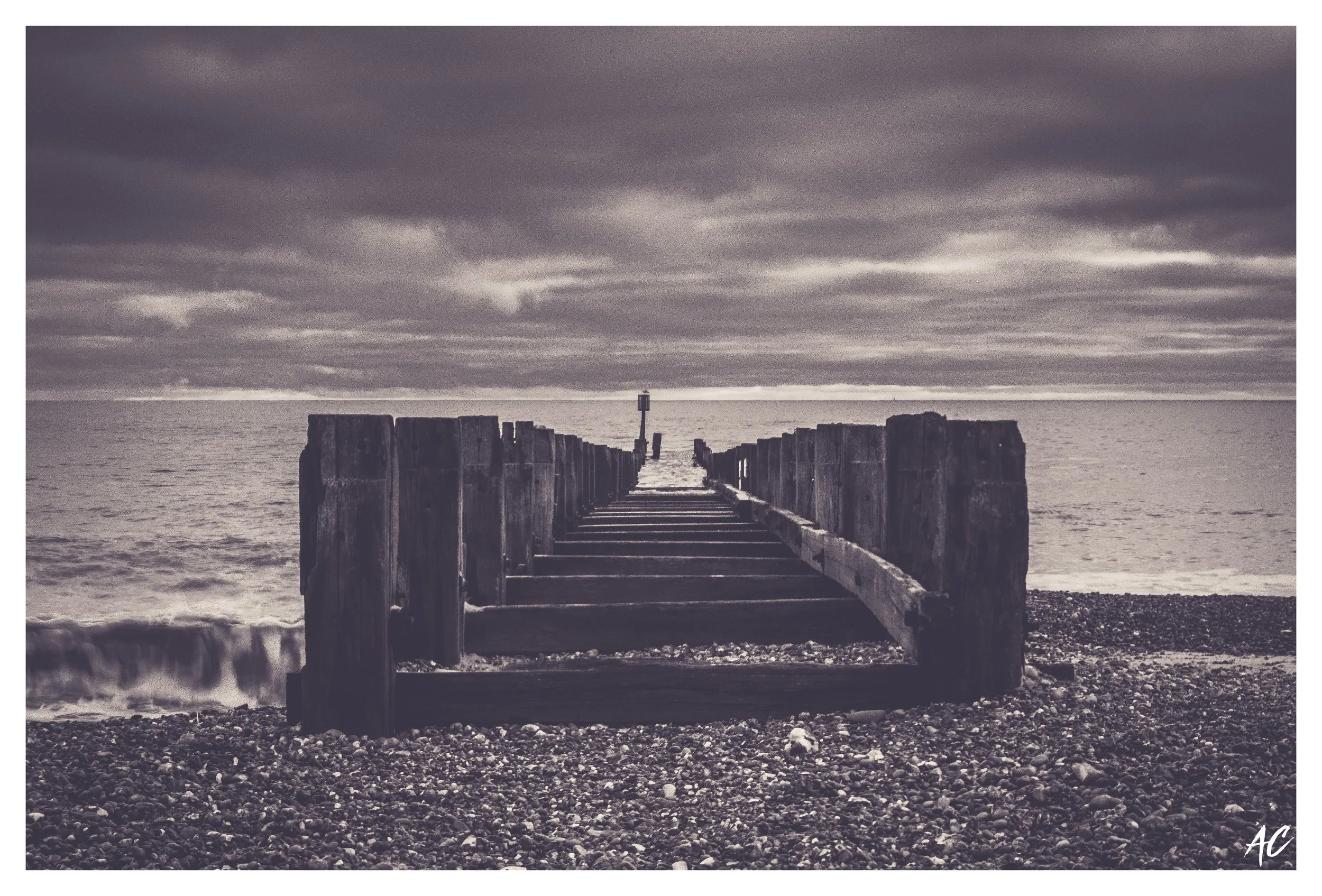 A black and white photograph of a wooden pier extending into the sea, with a cloudy sky overhead and a rocky beach in the foreground.