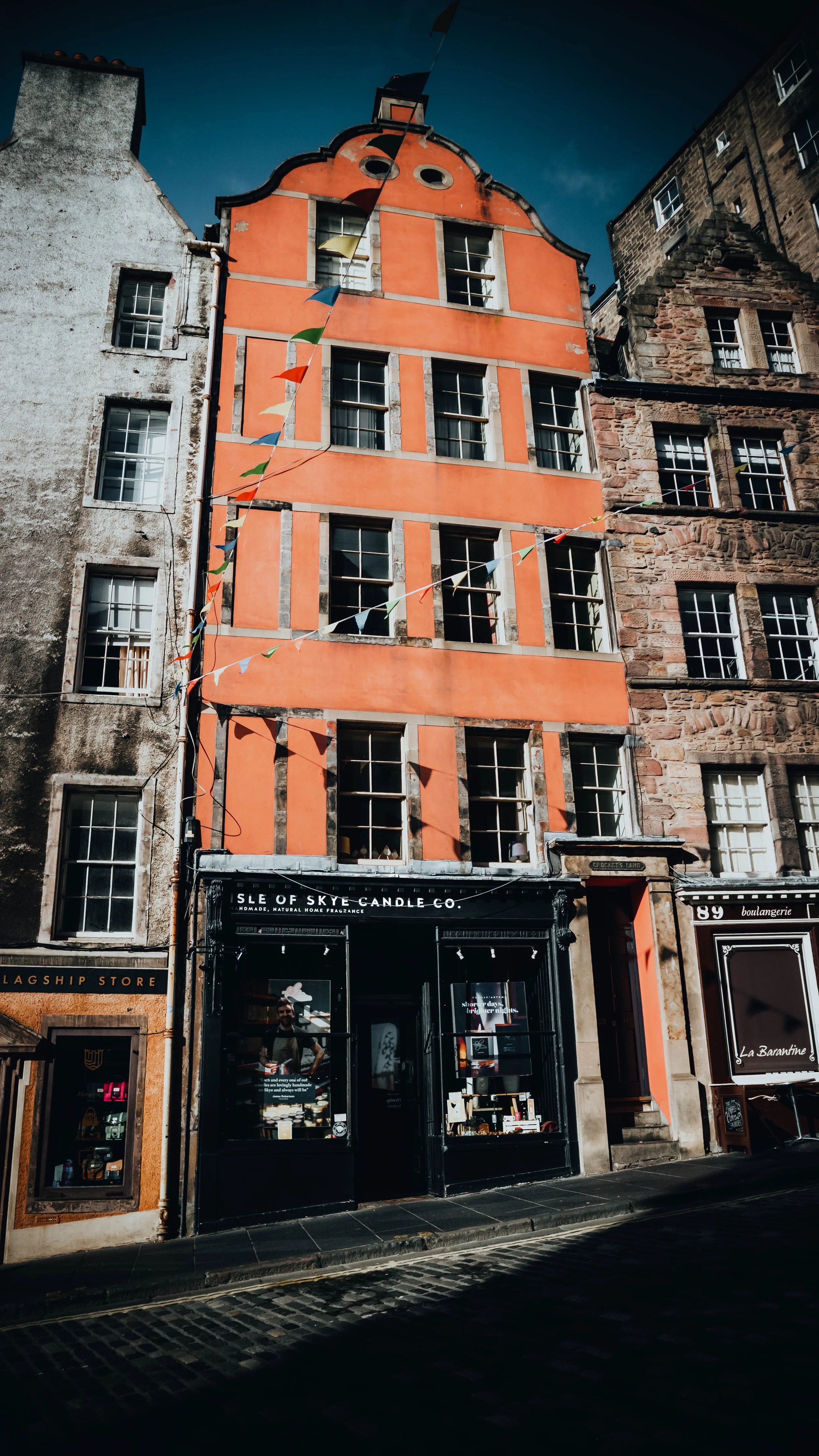 Colorful building with orange and gray facades, multiple windows, black storefront with white sign reads 'Isle of Skye Candle Co.' colorful flags hanging in front, neighboring buildings with stone and white facades, blue sky.