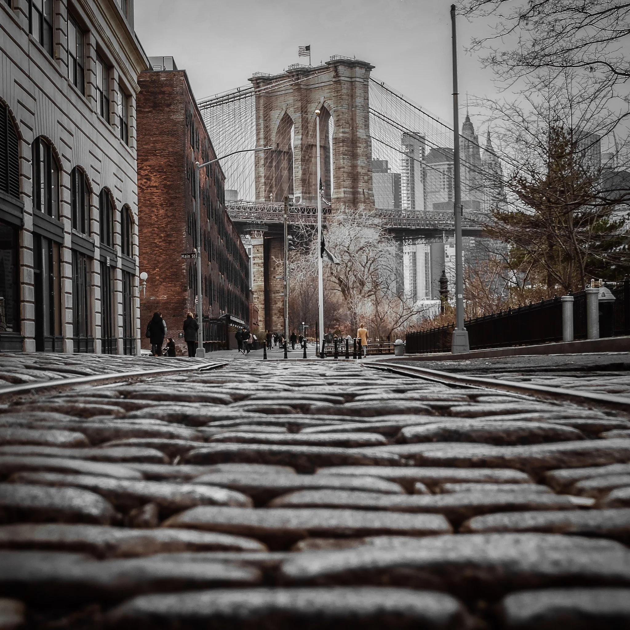 A low-angle view of cobblestone street with buildings on the left, trees on the right, and the Brooklyn Bridge in the background under cloudy sky.