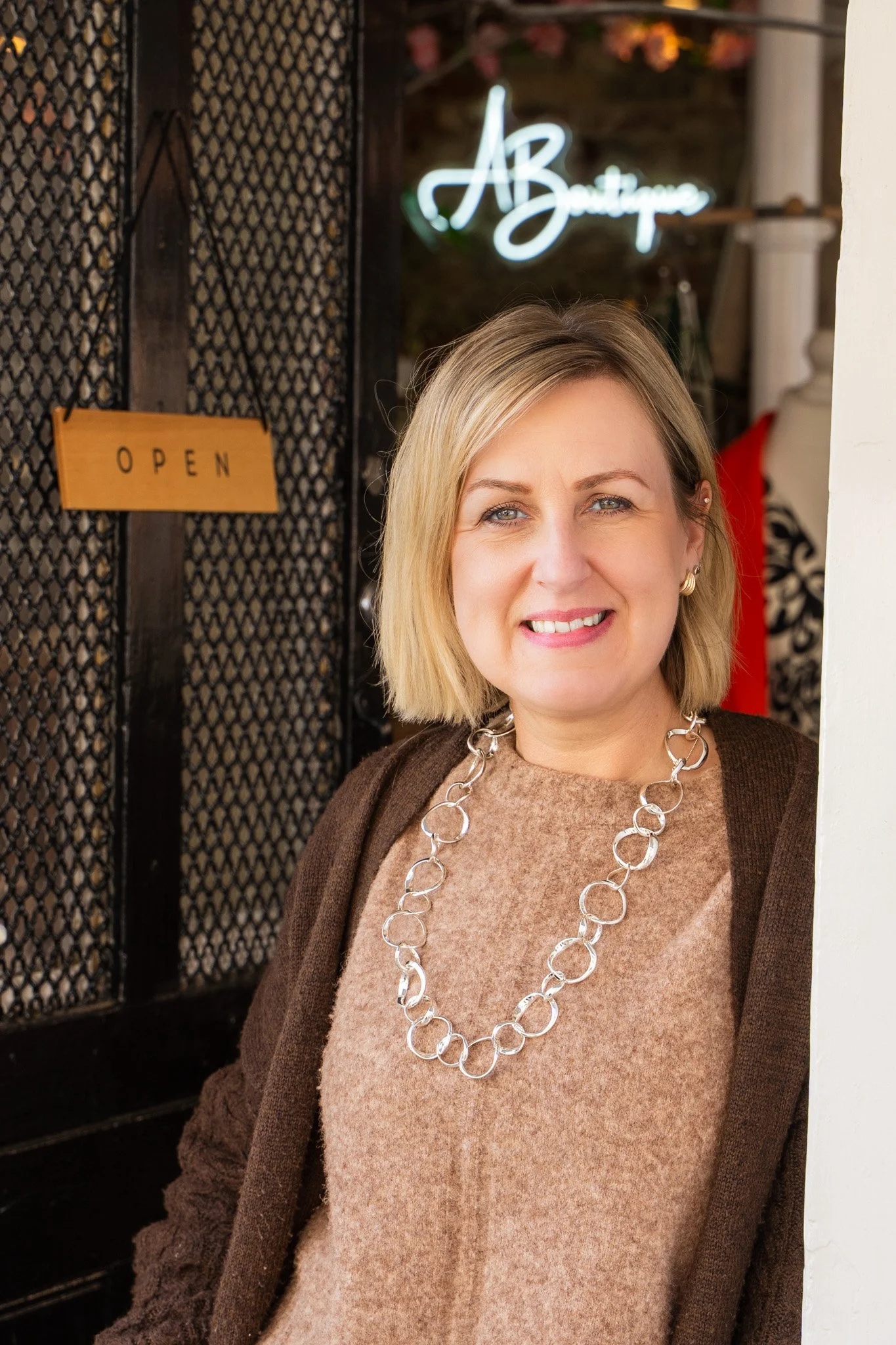 A woman with blonde hair and blue eyes standing inside a boutique, smiling at the camera. She wears a beige top, a brown cardigan, and a large silver chain necklace. Behind her, a black wire mesh partition features a small yellow 'OPEN' sign, and a n