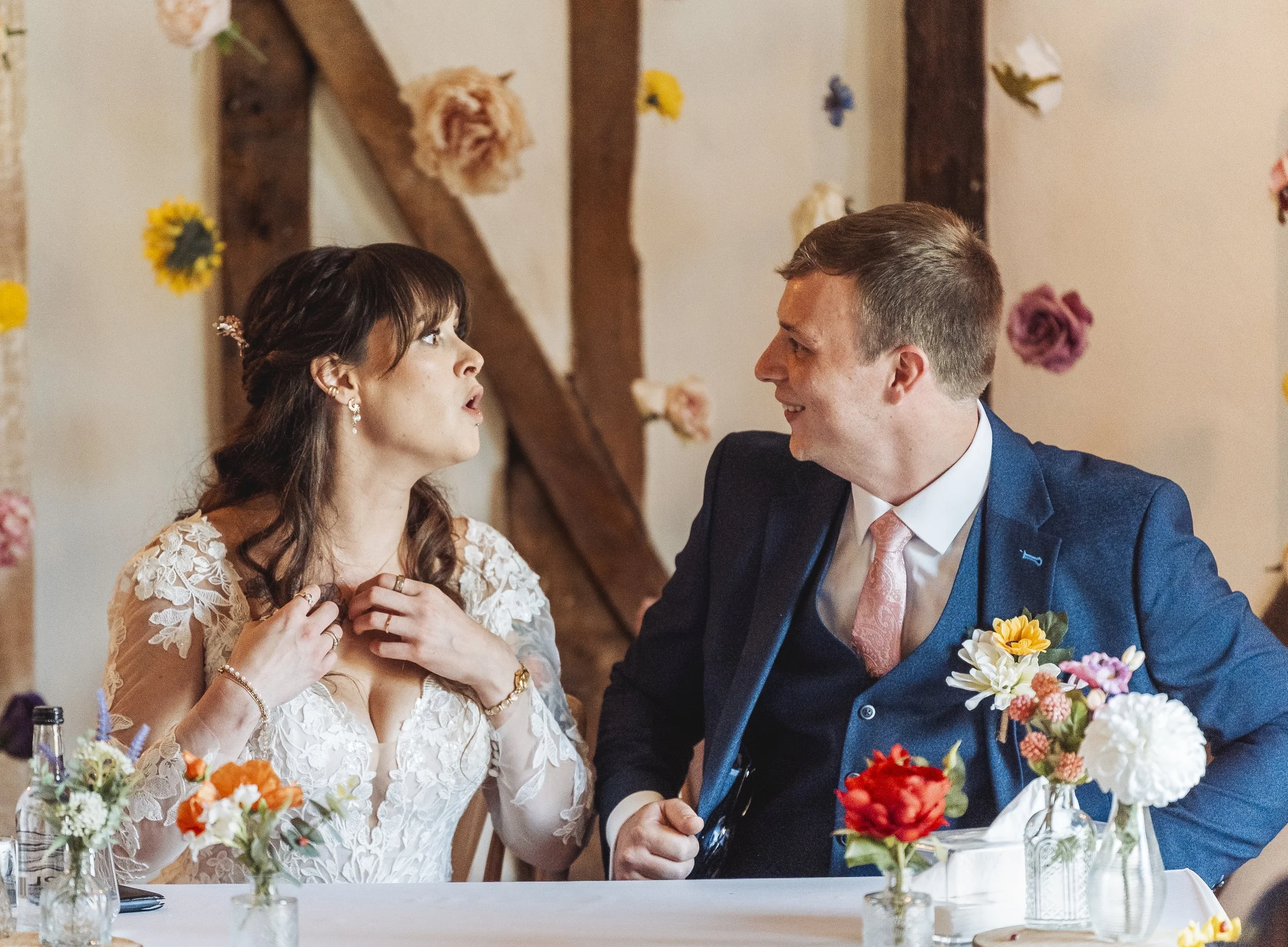 A bride and groom sitting at a wedding reception table, holding hands and looking at each other. The bride has dark hair, is wearing a lace wedding dress and jewelry. The groom has short hair, is wearing a navy blue suit with a pink tie. The backgrou