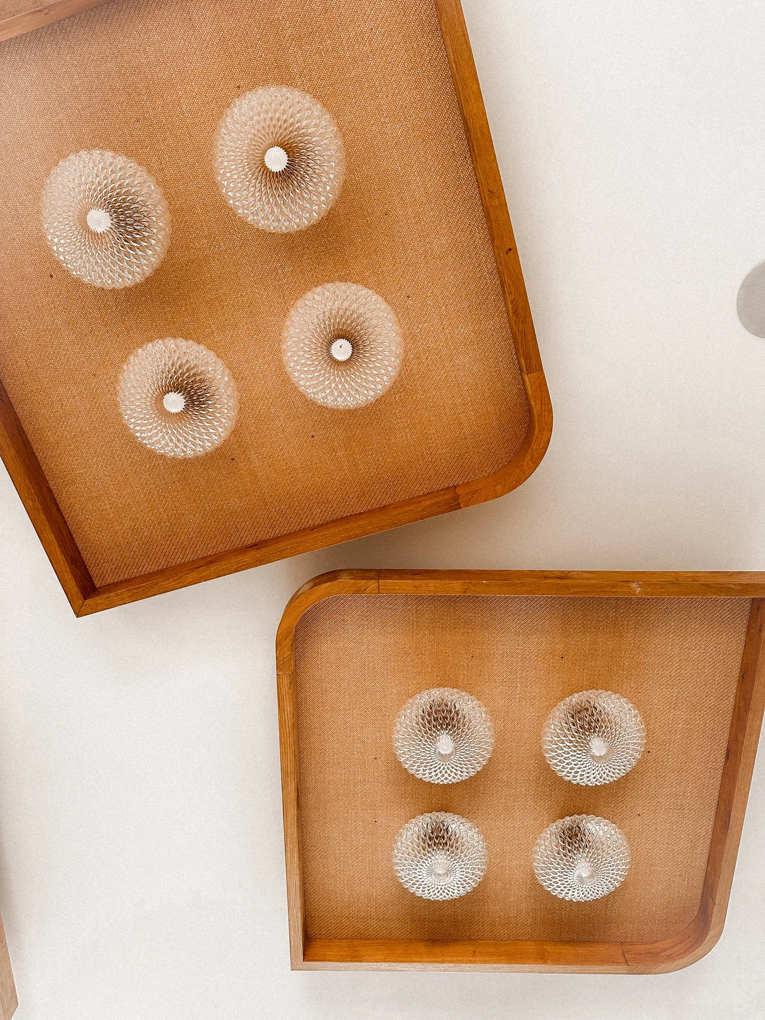 Two wooden trays with orange textured surfaces holding six glass candle holders with intricate designs, placed on a white surface.