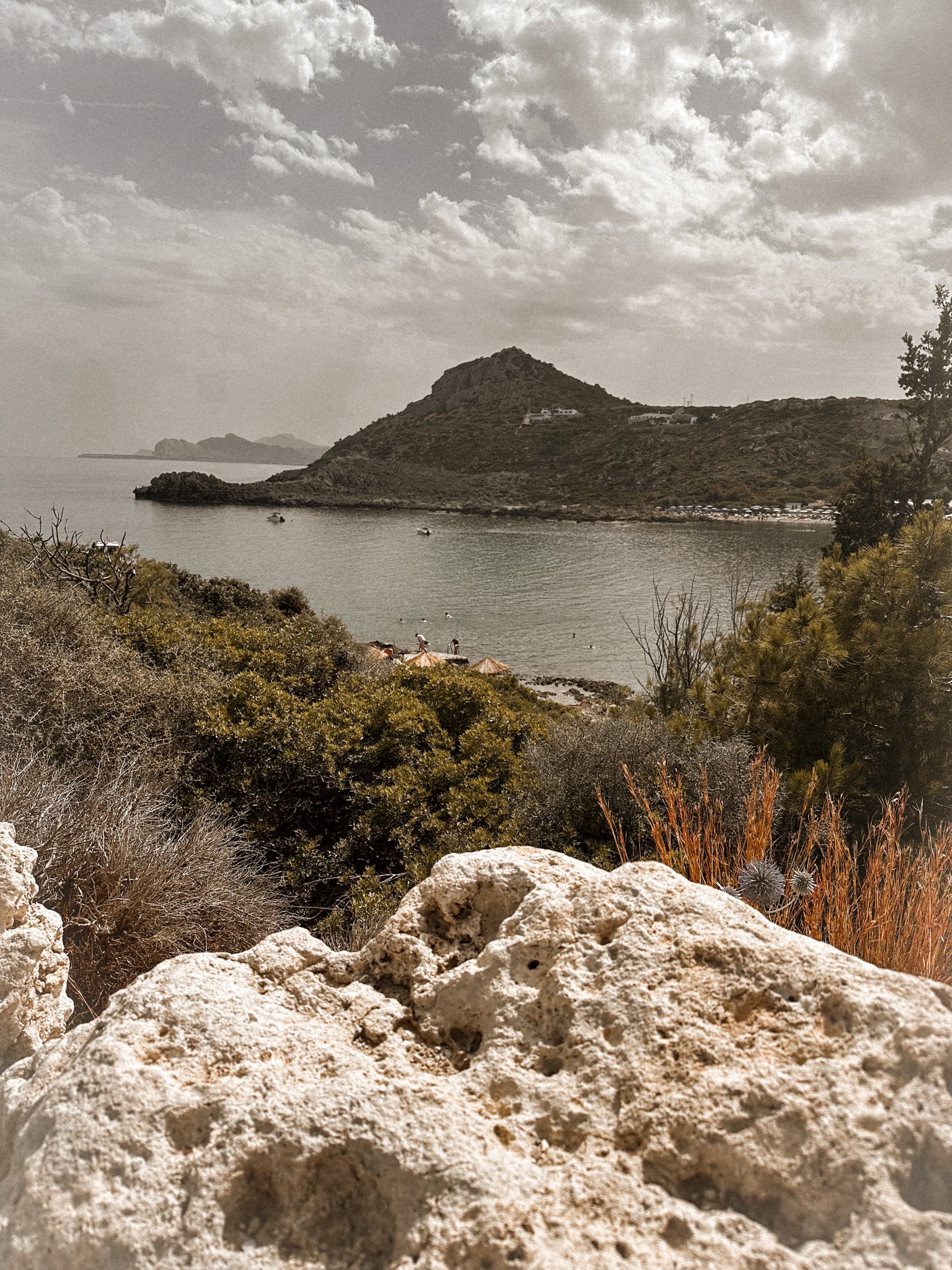 A scenic view of a bay with calm waters, a small boat, and rocky hills in the background. Vegetation and bushes are in the foreground, with a partly cloudy sky above.