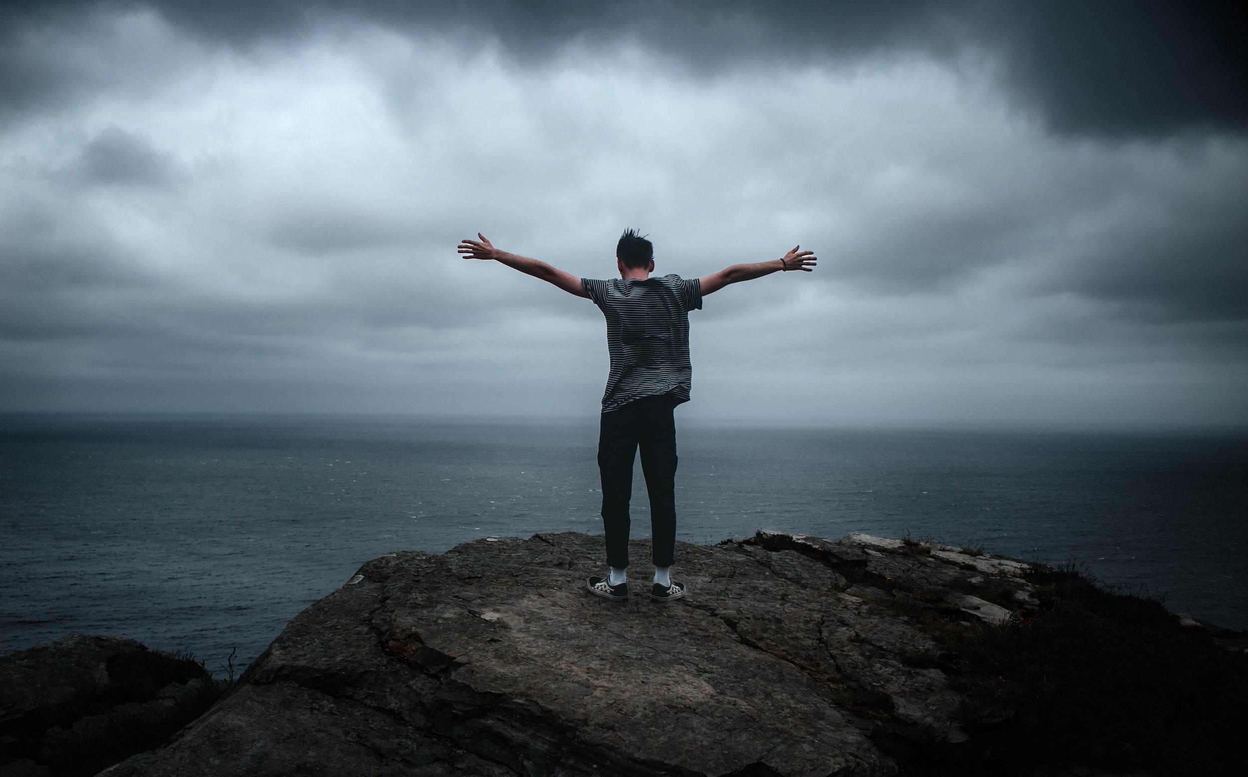 A person standing on a rocky cliff with arms outstretched, facing the ocean under cloudy skies.