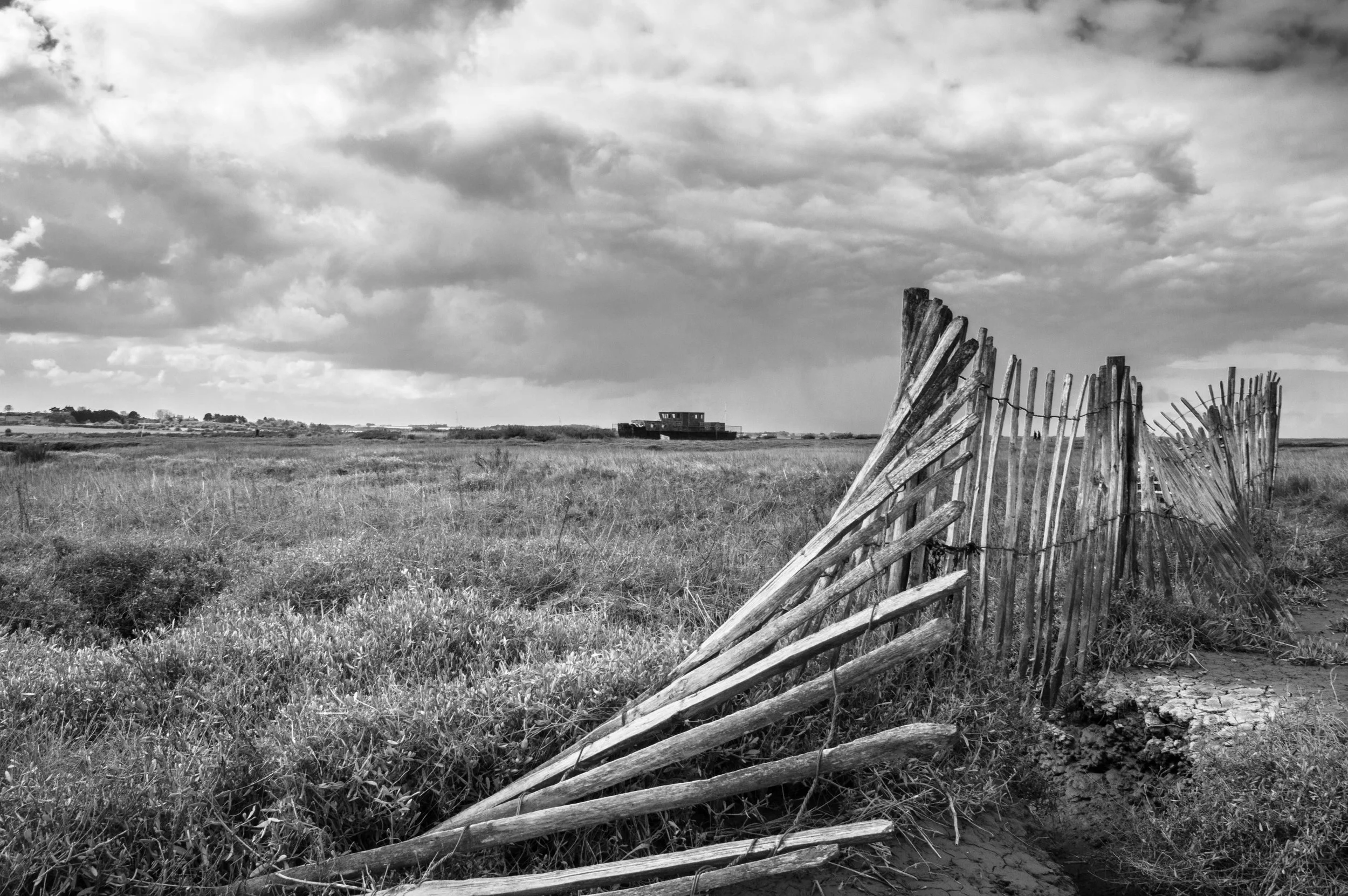 A black and white photo of a weathered wooden fence in a grassy field with structures in the background and a cloudy sky overhead.