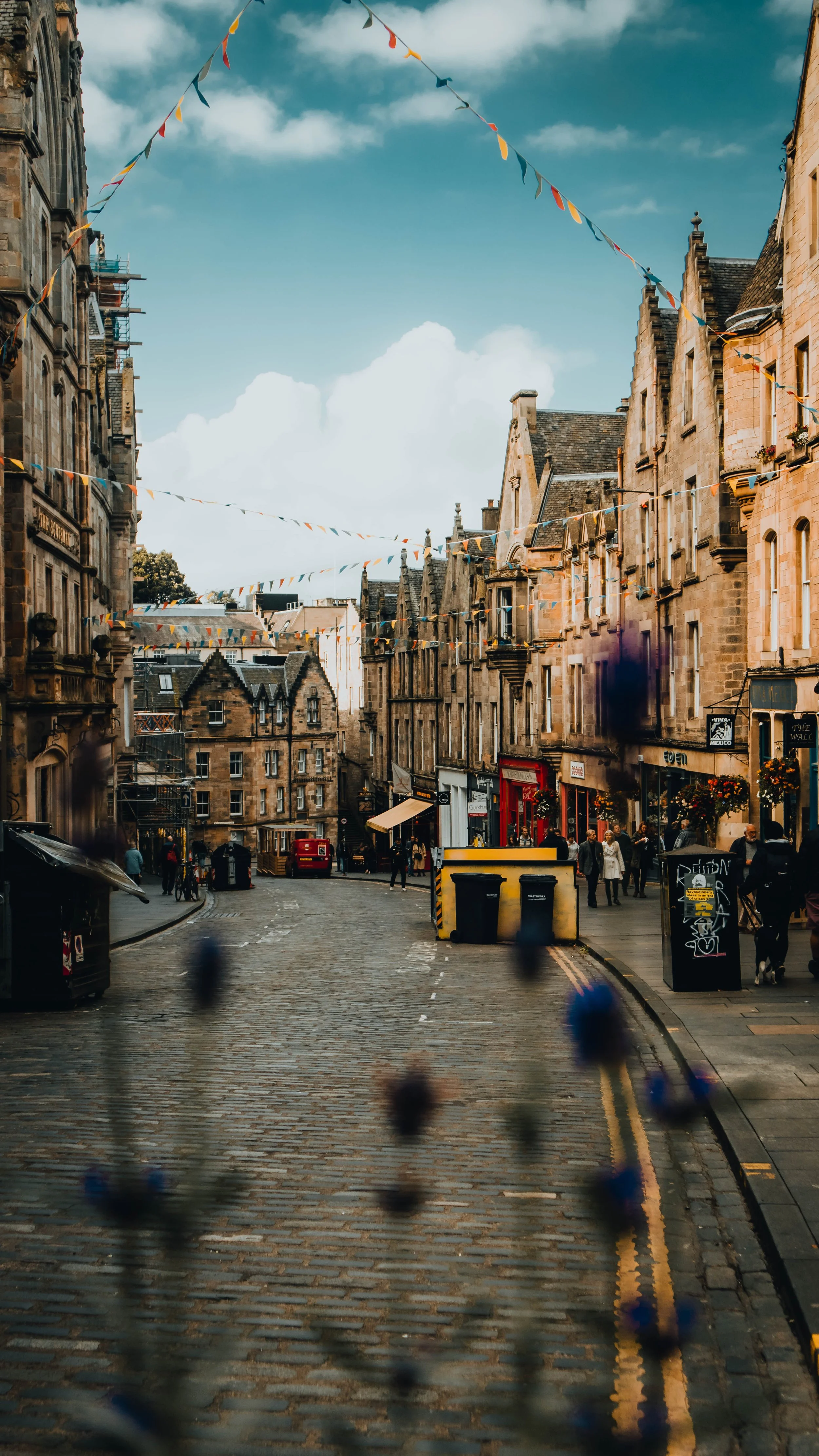 A street scene in a historic European city with cobblestone streets, old stone buildings, and decorative bunting hanging across the street. Pedestrians are walking along the sidewalks, some shops and cafes are visible, and the sky is partly cloudy.