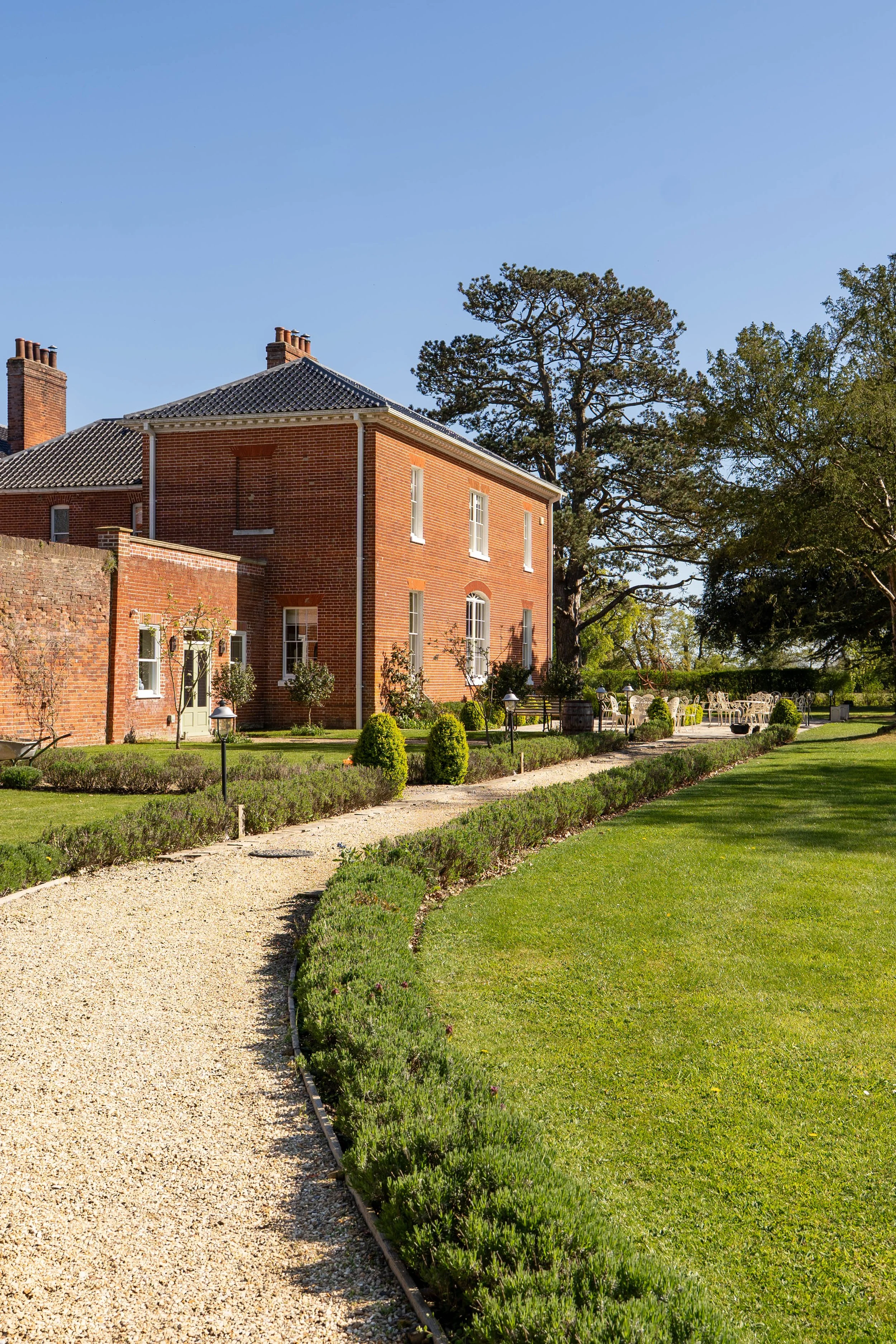 A brick house with a dark tiled roof and white window frames, surrounded by green trees and a neatly landscaped garden with a gravel pathway, grass, and trimmed bushes.