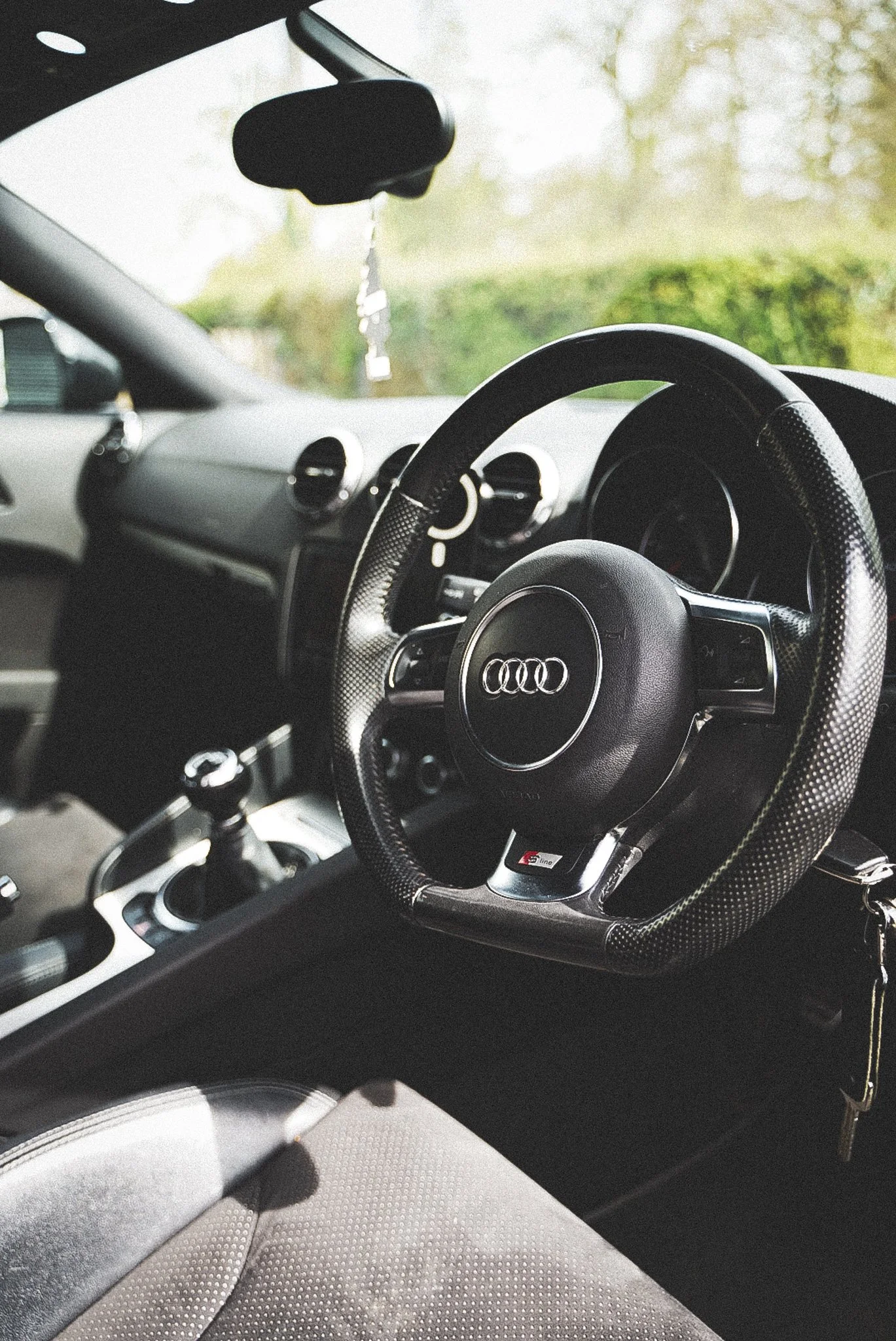 Inside view of a car showing the steering wheel with an Audi logo, dashboard, and part of the gear shift, with a blurred background of greenery outside the car window.