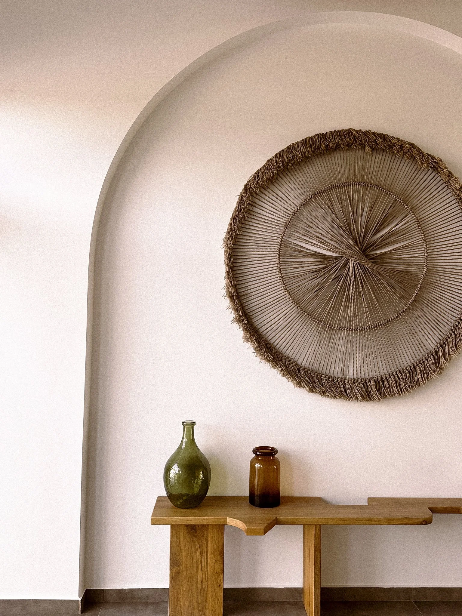 A wooden console table with three decorative vases (green, brown, amber) against a white wall. Above the table is a circular woven wall hanging with a fringed edge.