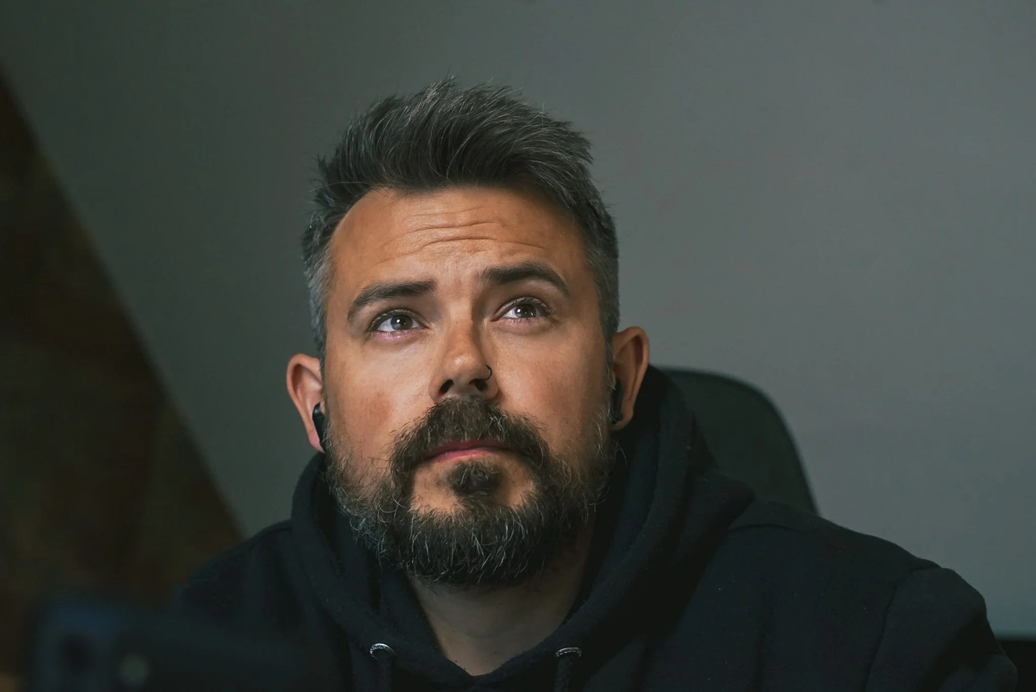 A man with dark hair and a beard, looking upwards with a serious expression, in an indoor setting.