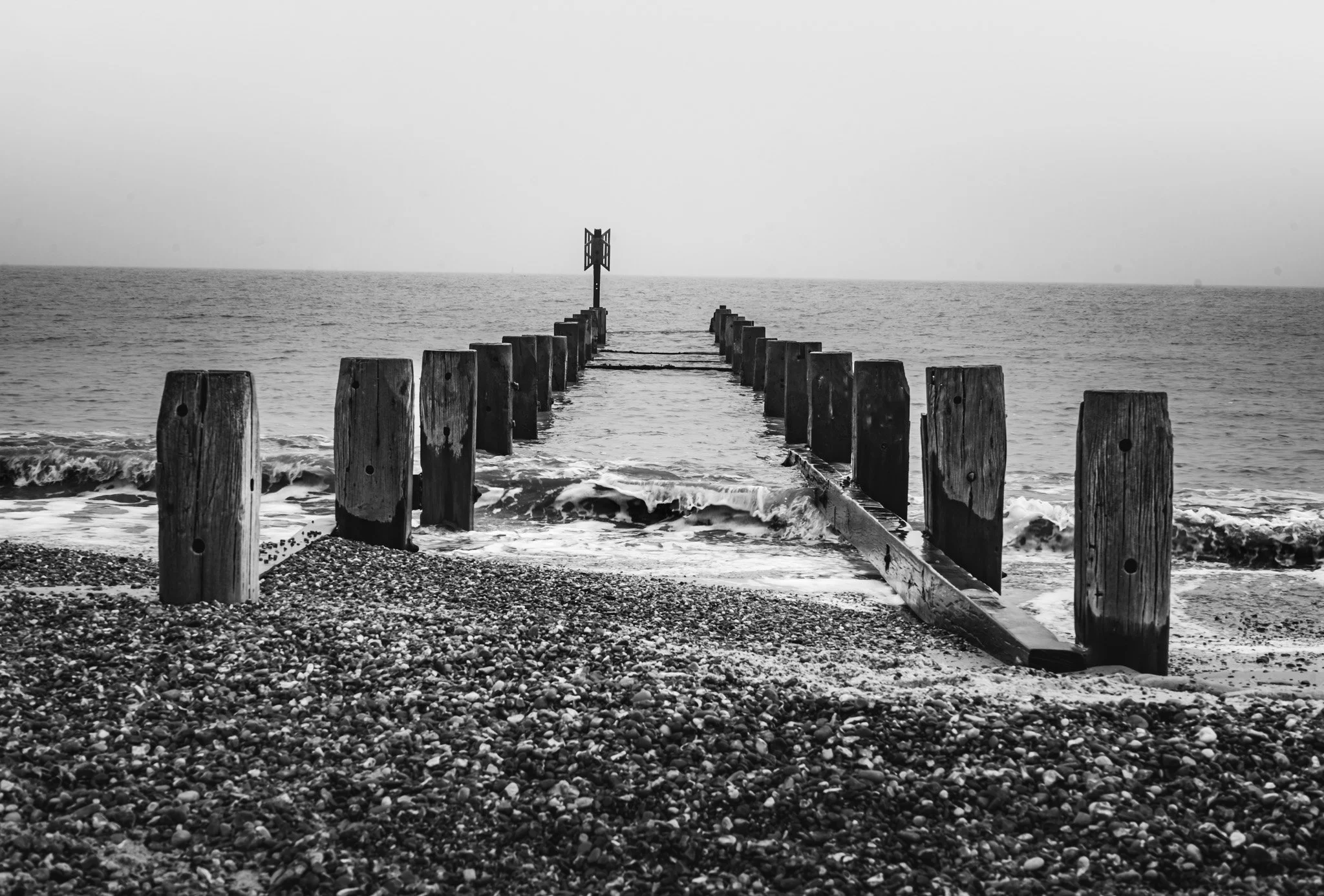 A black and white photo of a breakwater extending into the ocean with wooden posts and a small navigation sign at the end, waves crashing at the shoreline, and a cloudy sky.