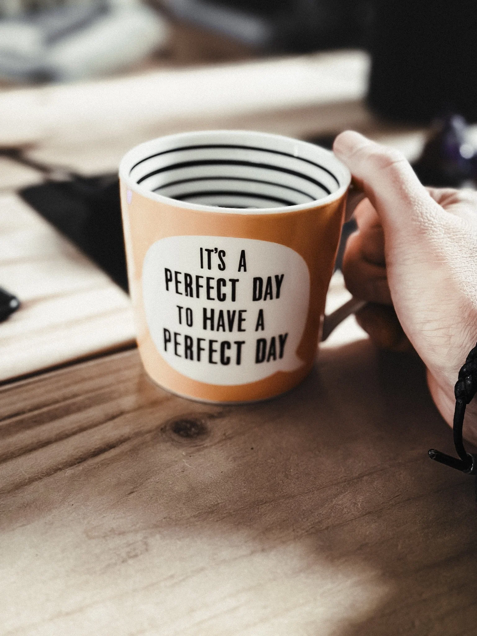 A person holding a coffee mug with the text 'It's a perfect day to have a perfect day' written on it. The mug is on a wooden table with some blurred objects in the background.