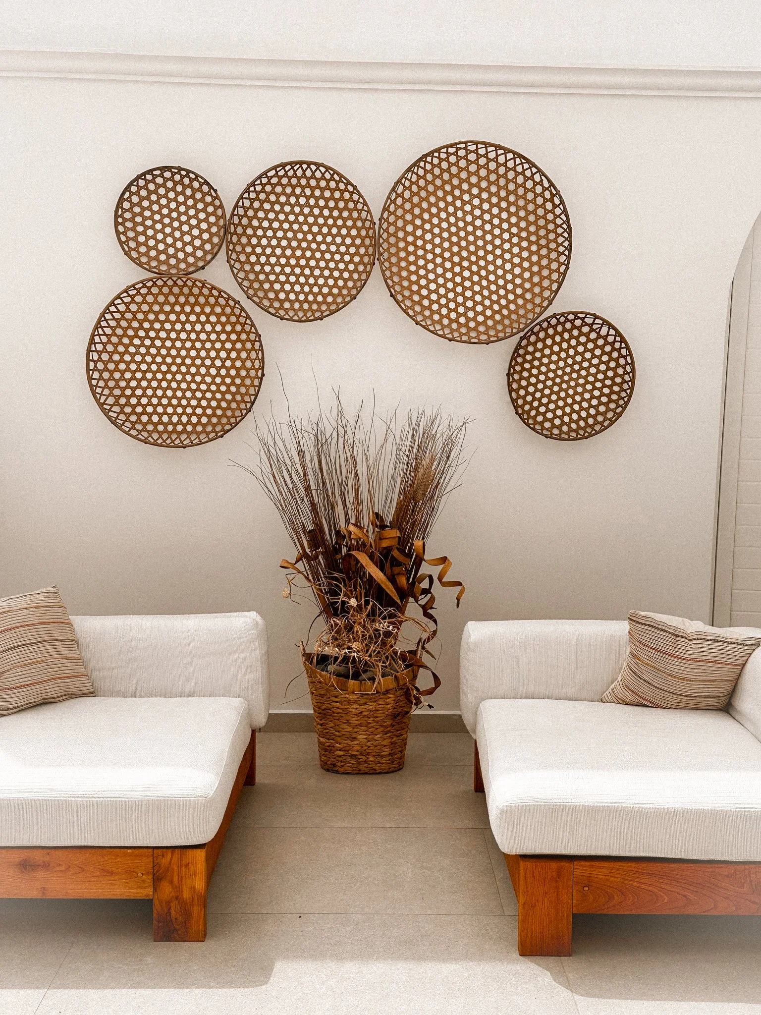 Two white sofas with wooden bases facing each other, separated by a large woven basket containing dried grass and decorative ribbons. Behind the basket, six round wicker wall decorations of varying sizes are hung on a white wall.