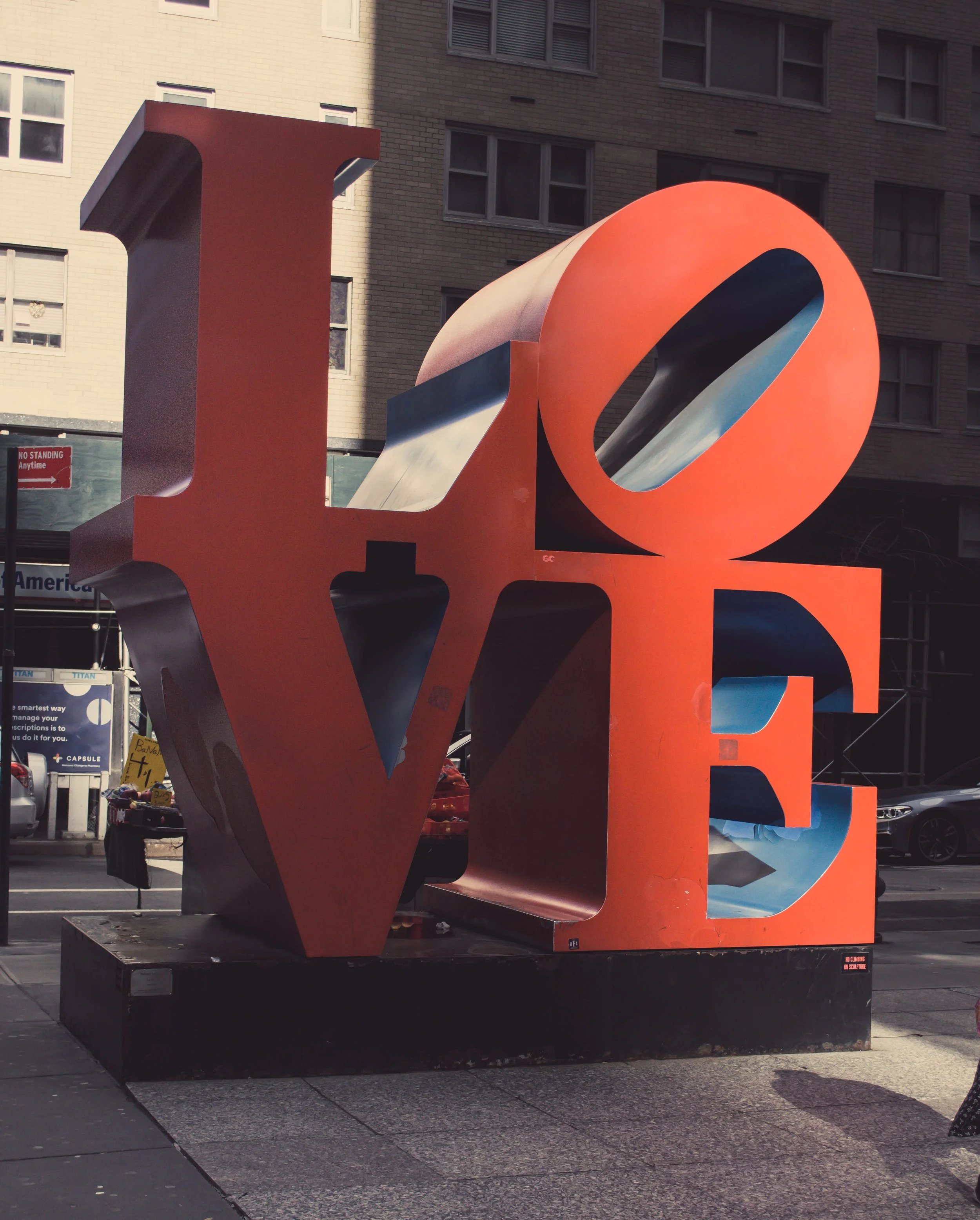 Large colorful sculpture of the word "LOVE" with the letter "L" and the letter "O" shown, set outdoor in front of a building with multiple windows.