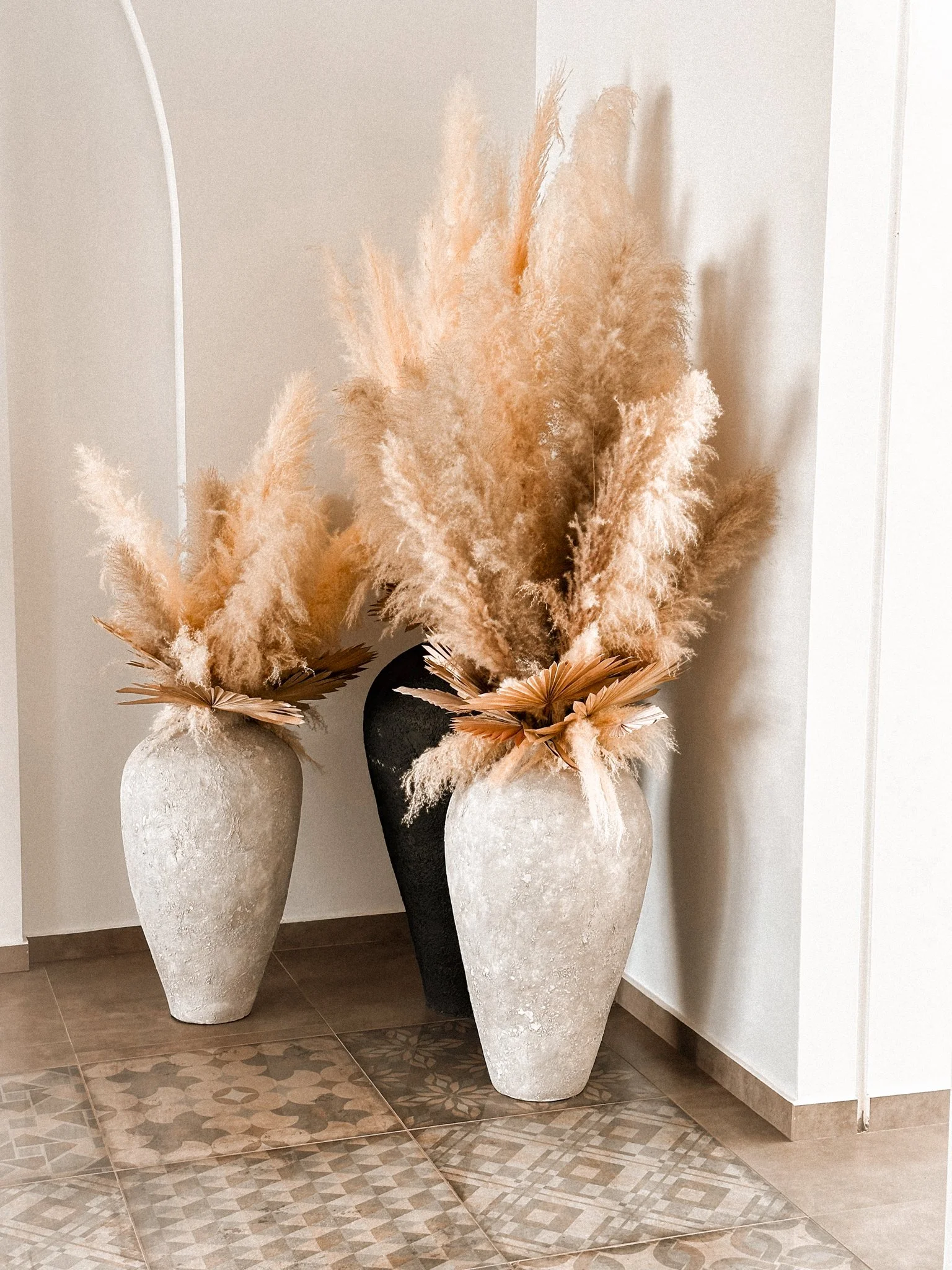 Three large decorative vases with dried pampas grass and palm leaves arranged inside them, placed on a tiled floor against a beige wall.