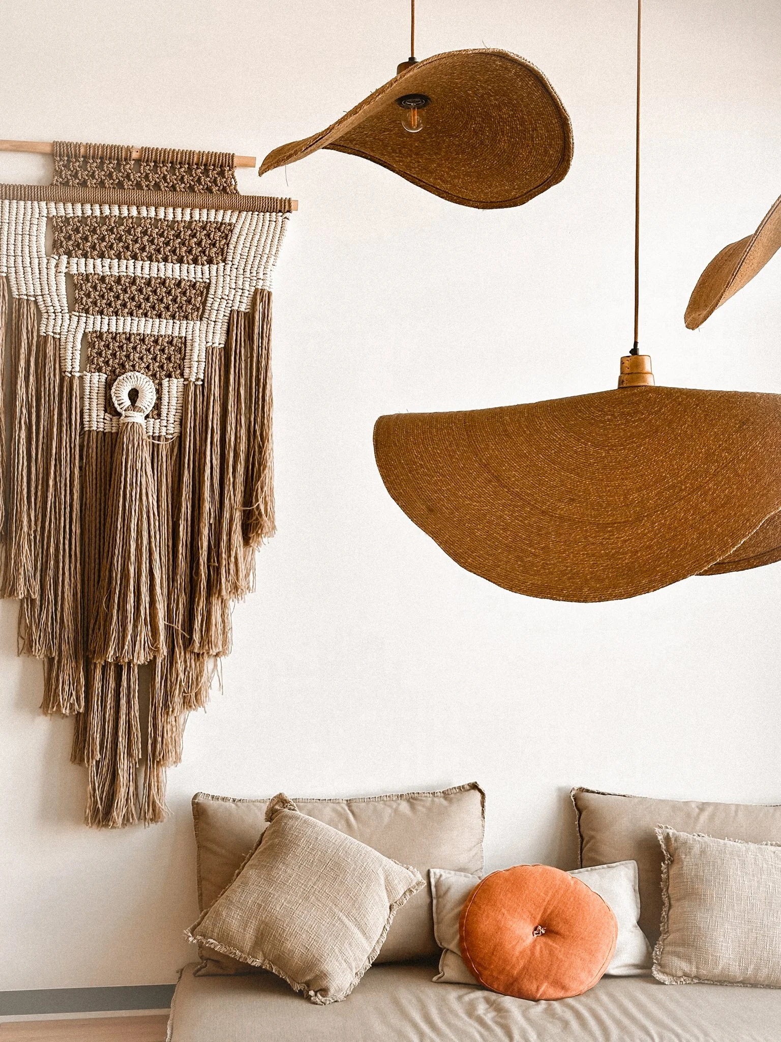 Living room wall decorated with woven wall hanging, hanging lamps, and pillows on a beige sofa.