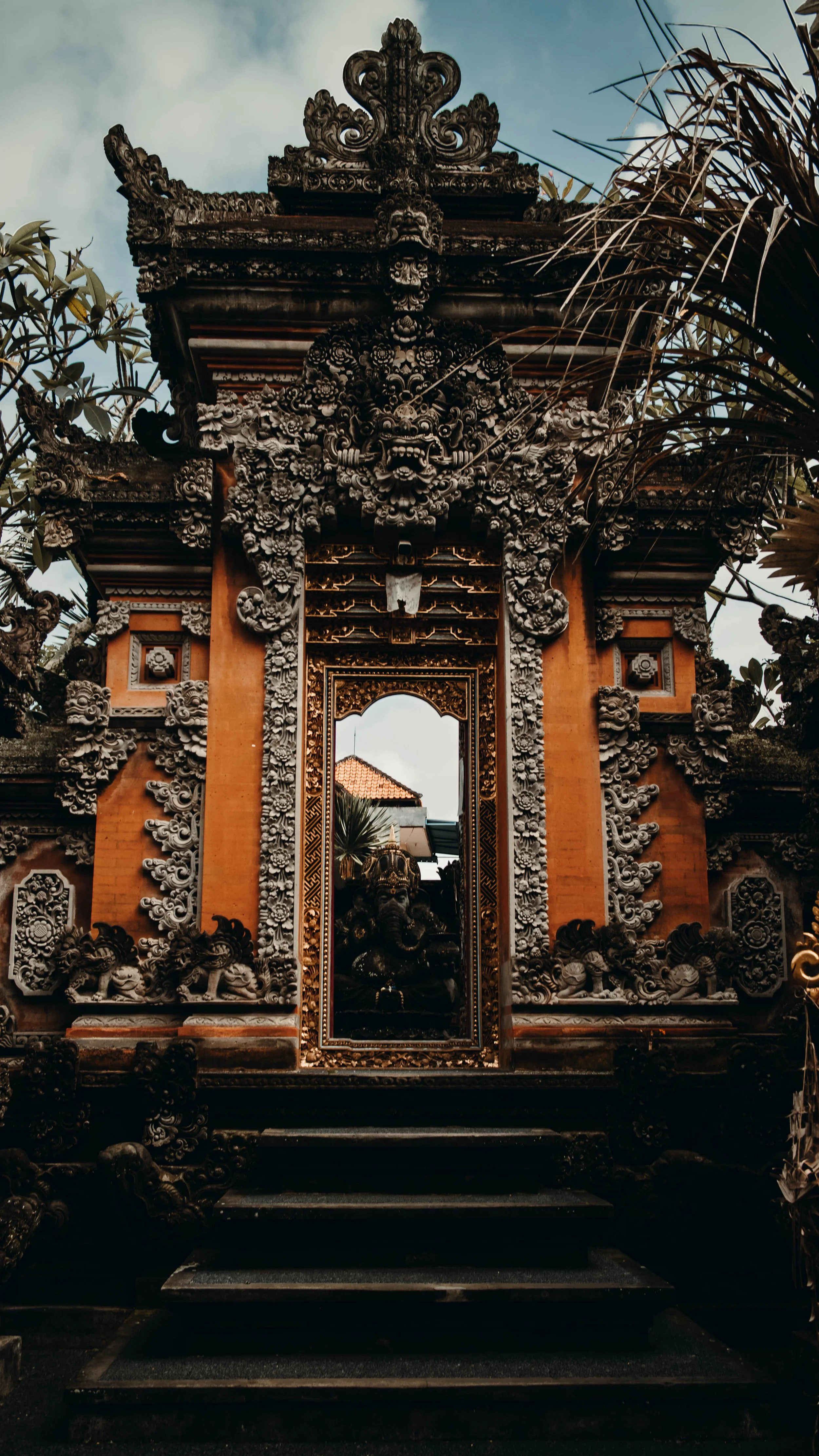 Ornate Balinese temple gate with detailed carvings, a statue of Ganesha inside, surrounded by trees and a cloudy sky.