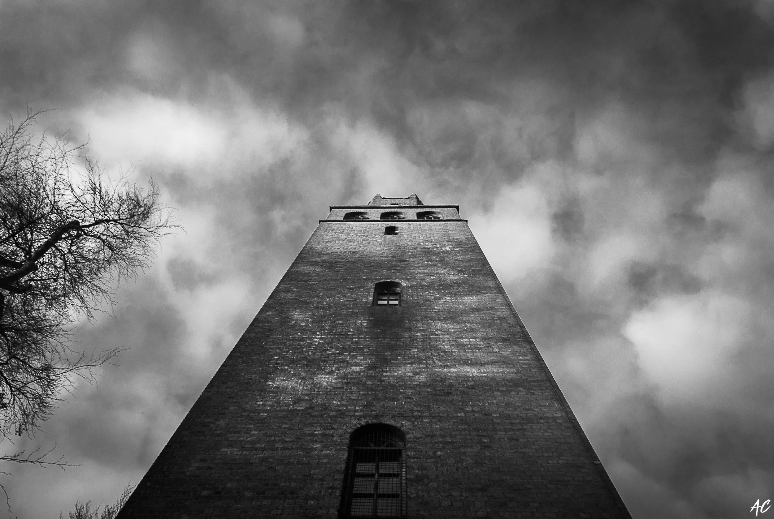 Black and white photo of a tall brick tower viewed from the base, with clouds and a leafless tree in the background.