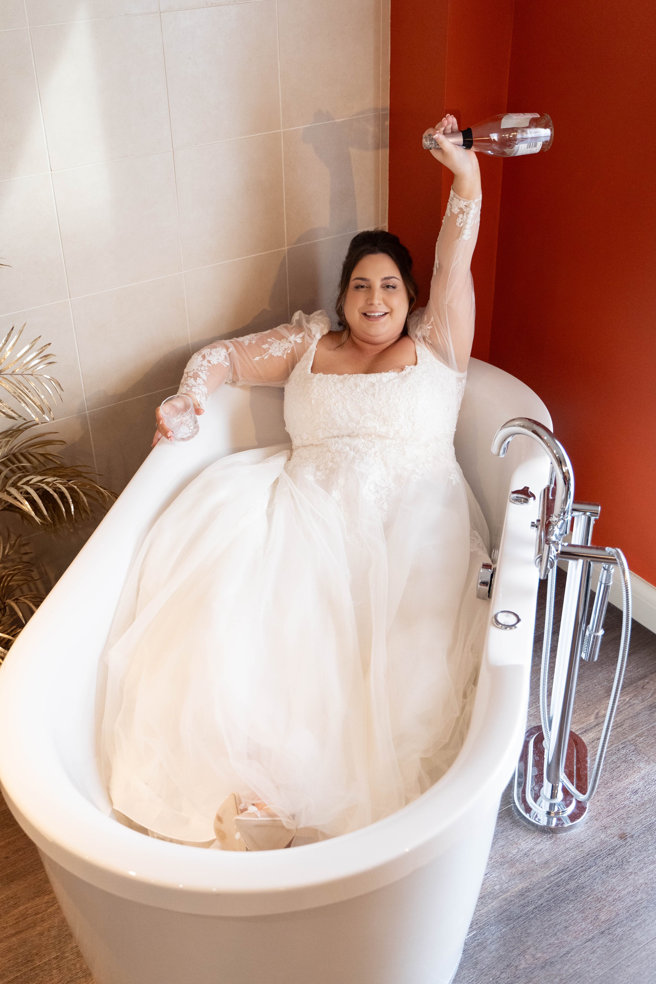 A woman in a wedding dress sitting in a bathtub, holding a wine bottle up in one hand and a glass in the other, with a smile on her face.