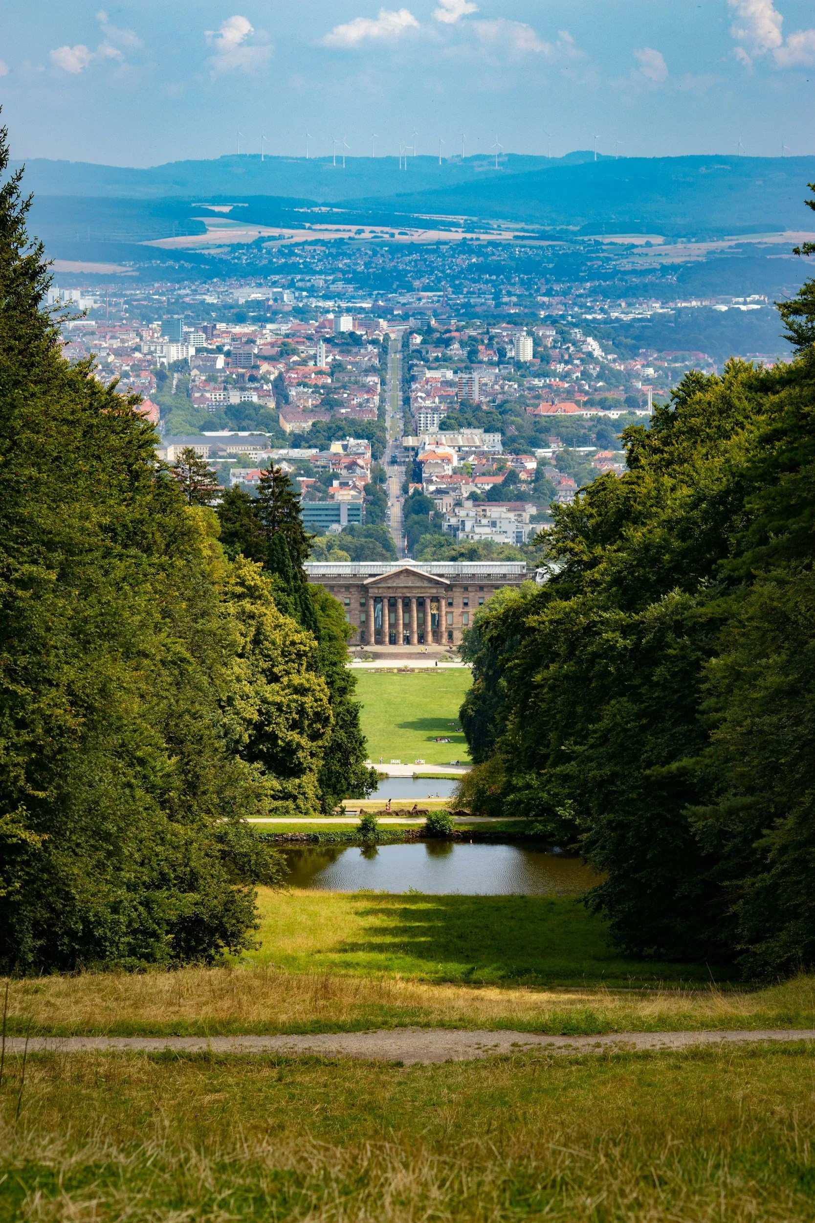 Blick durch einen Park auf eine Stadt mit Gebäuden, Bäumen, einem Wasserbecken und Hügel im Hintergrund, auf denen Windkraftanlagen stehen.