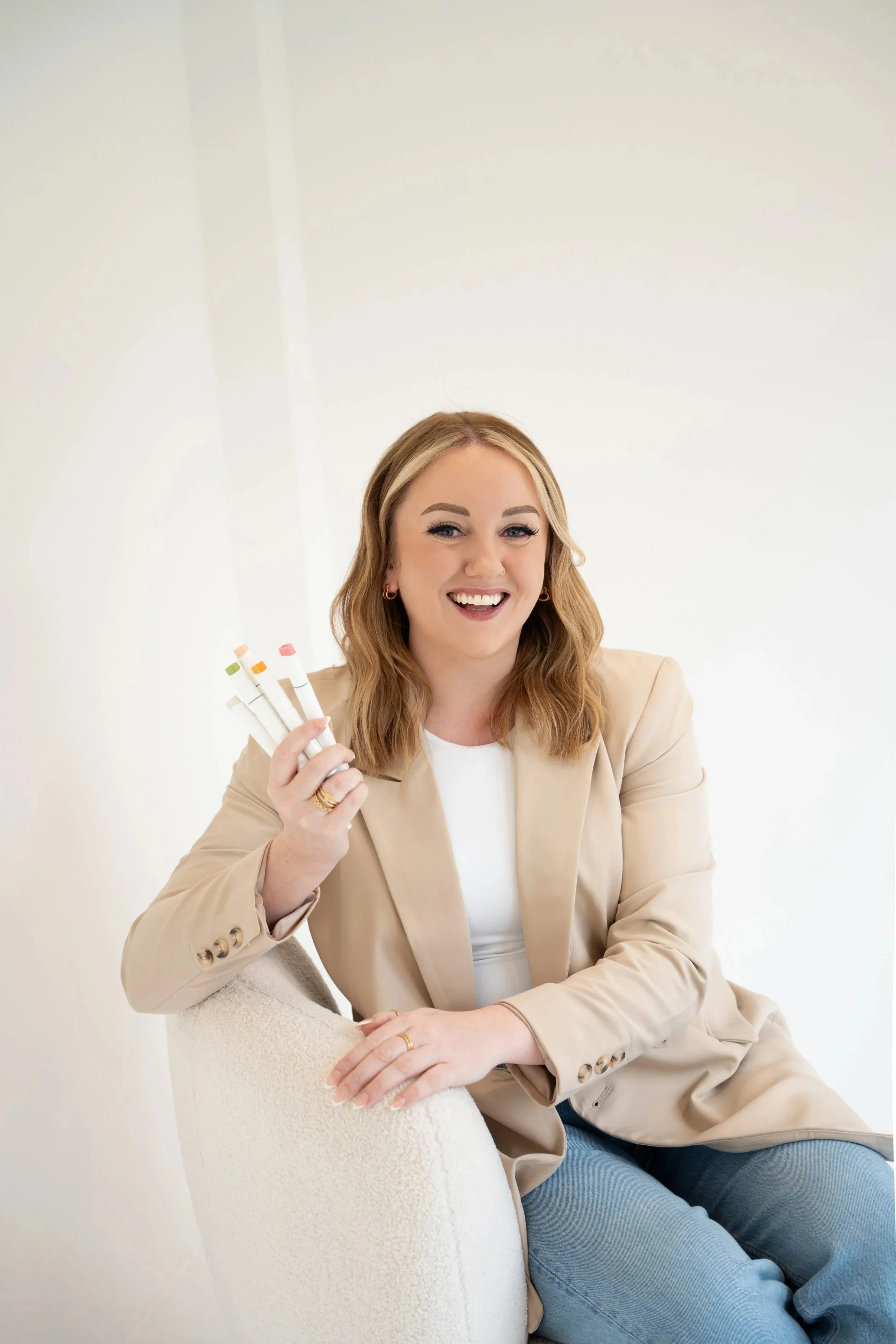 A woman with blonde hair, smiling and holding color swatches, sitting in a beige blazer and light-colored top.