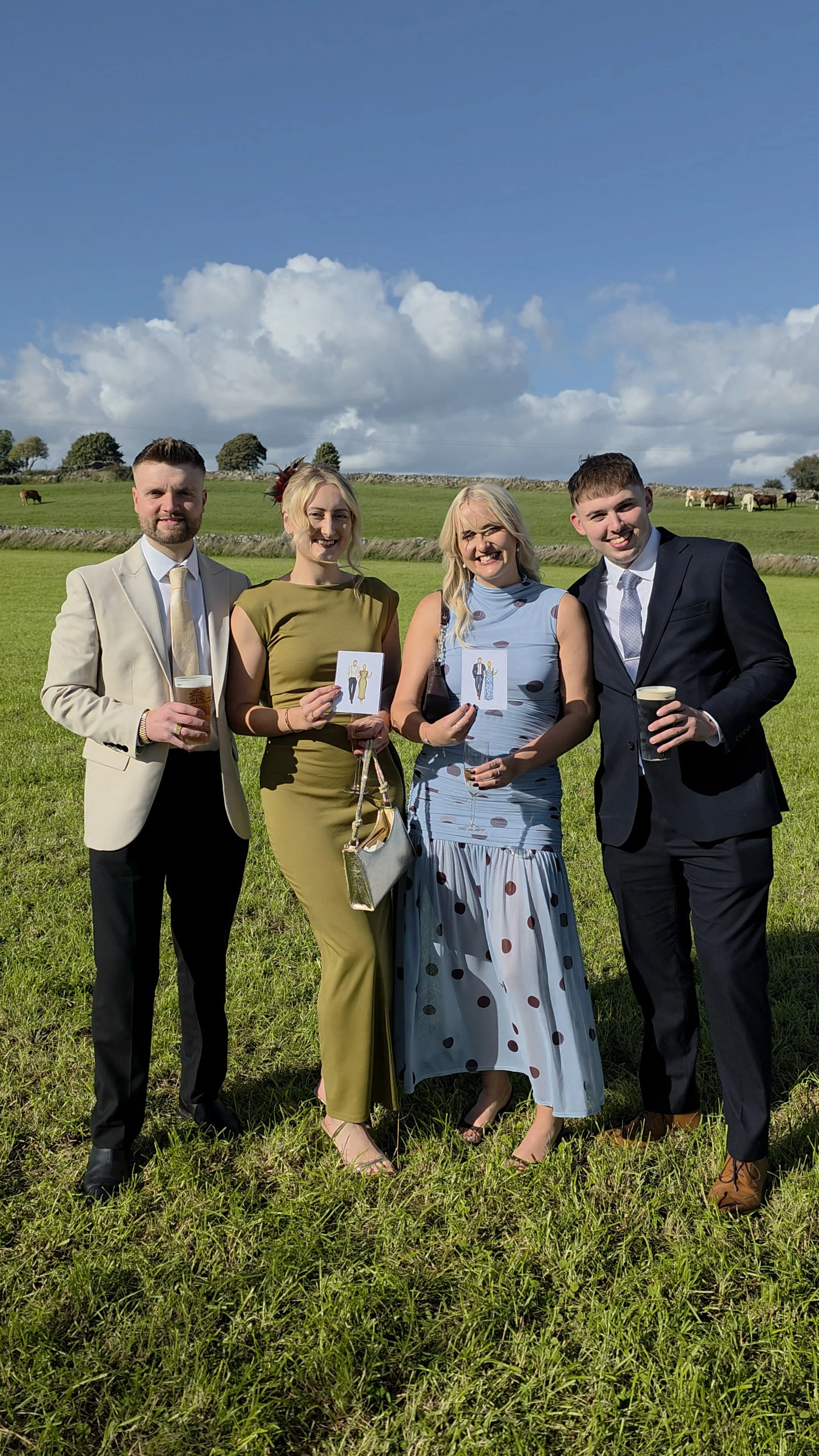 Four people dressed in formal attire standing outdoors on grass with a backdrop of rolling fields, horses, and a partly cloudy sky.