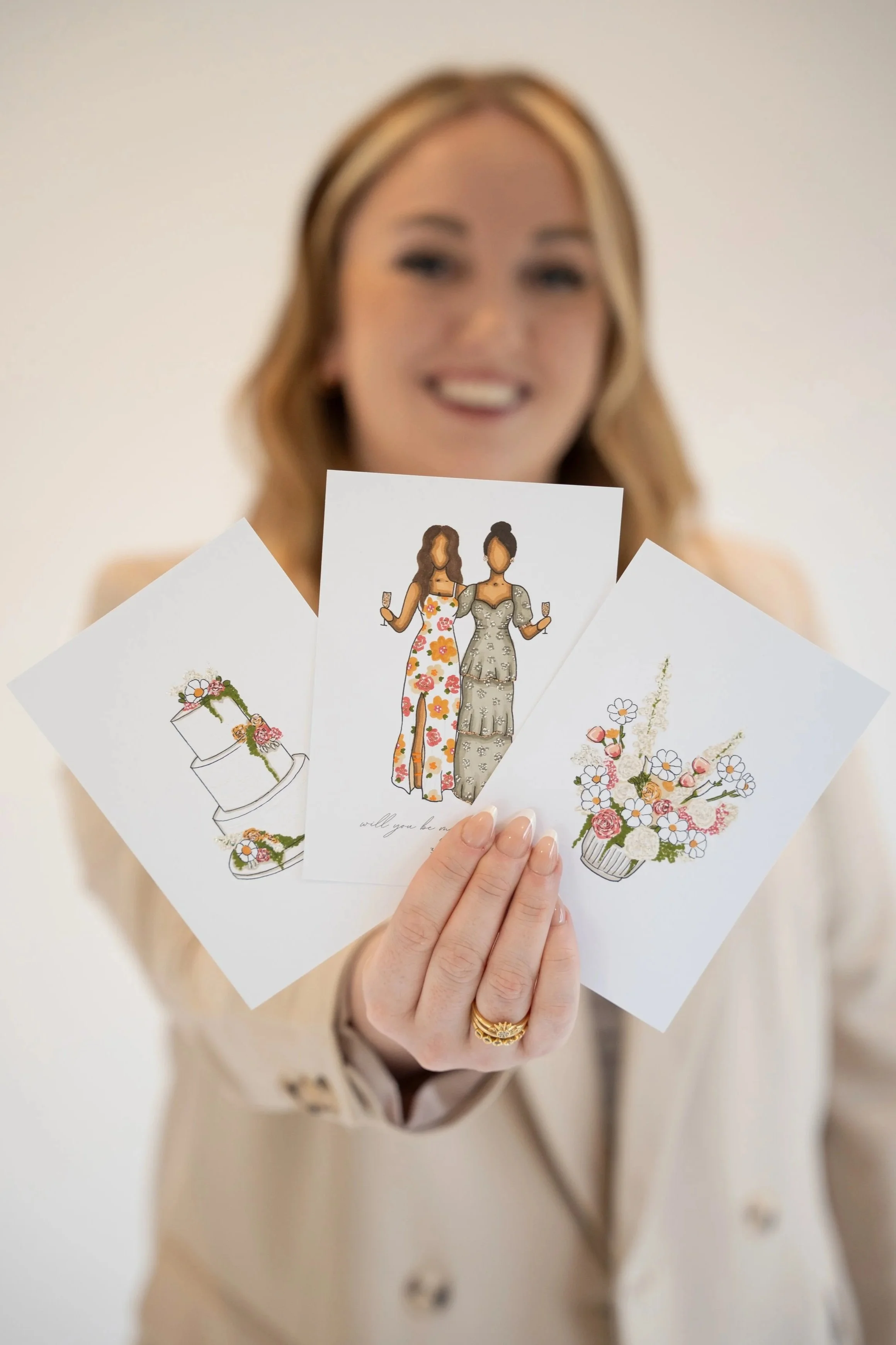 A woman holding three greeting cards with illustrations of women in dresses holding glasses of champagne, with a white background.