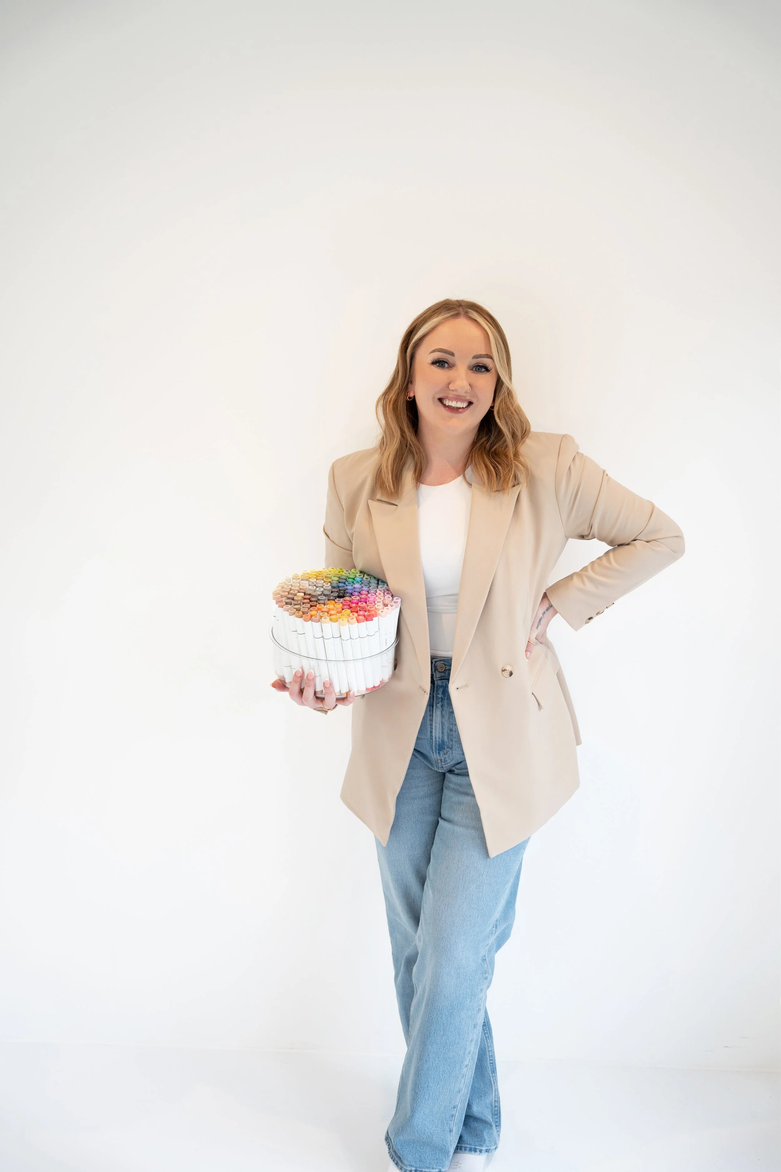 A woman with blonde hair, wearing a beige blazer and light blue jeans, standing against a plain white background, holding a large container filled with colorful markers, smiling at the camera.