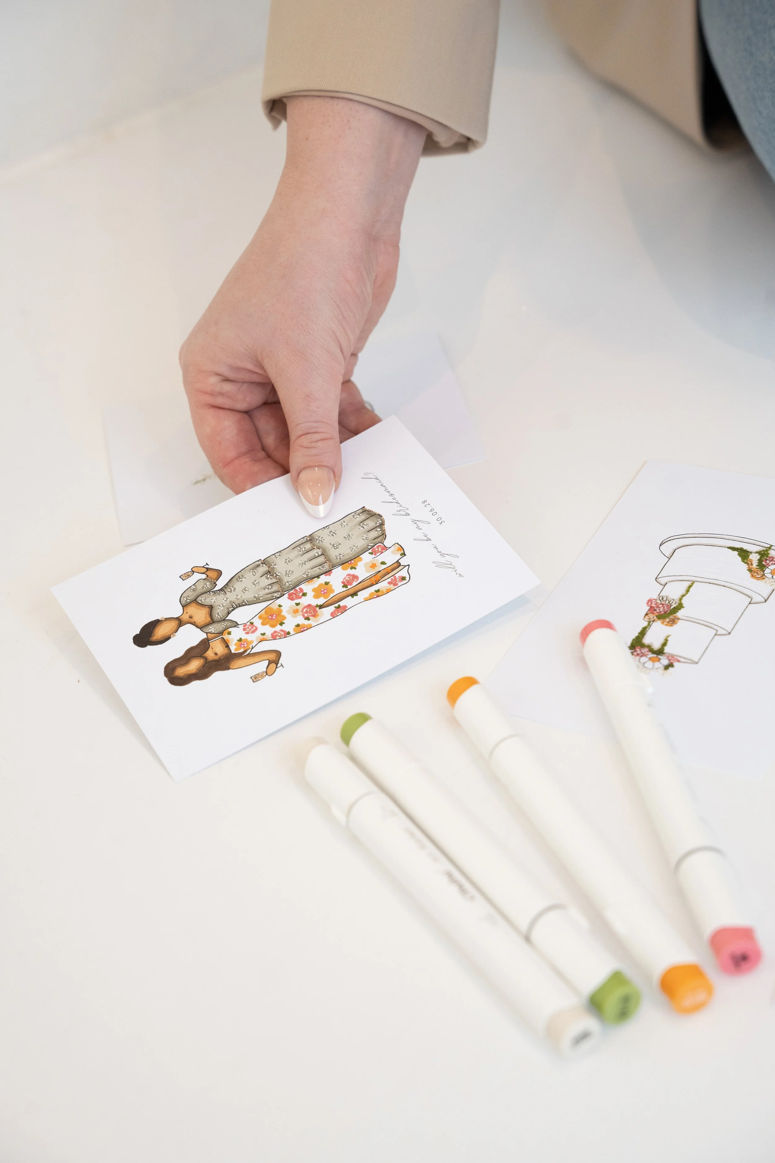 A hand holding a greeting card with a dog illustration, surrounded by white markers with colored caps, on a white surface.