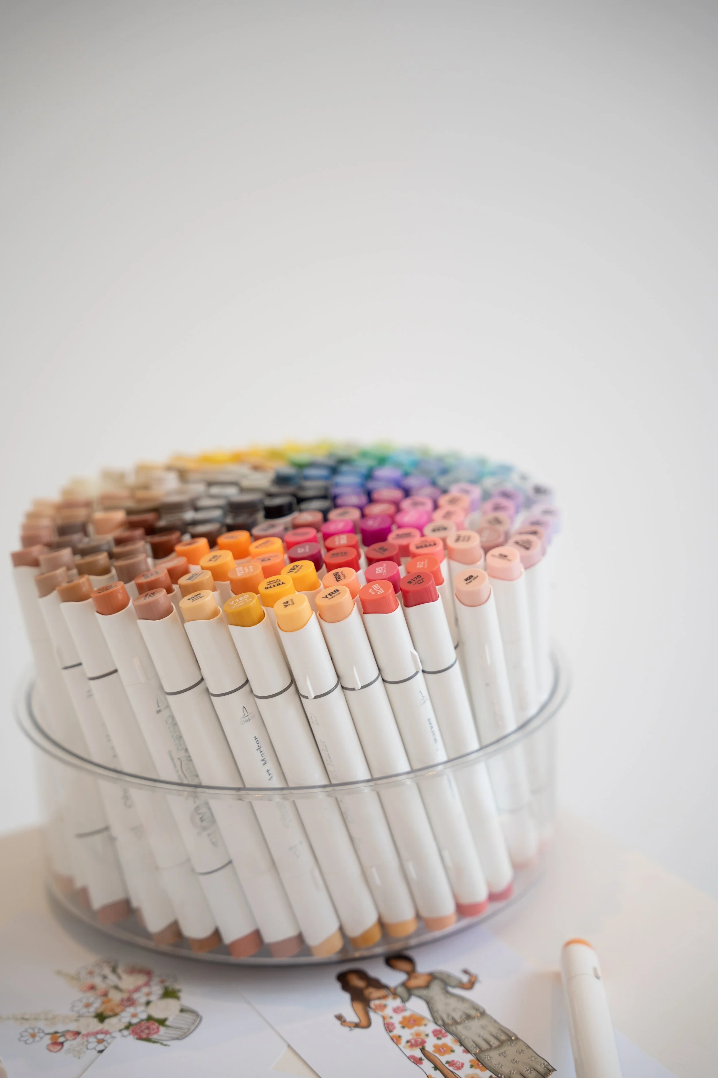 A circular container filled with numerous colorful markers arranged in a gradient. Below the container are illustrated paper cutouts of women in floral dresses, one holding a bouquet, with a silver ruler nearby.