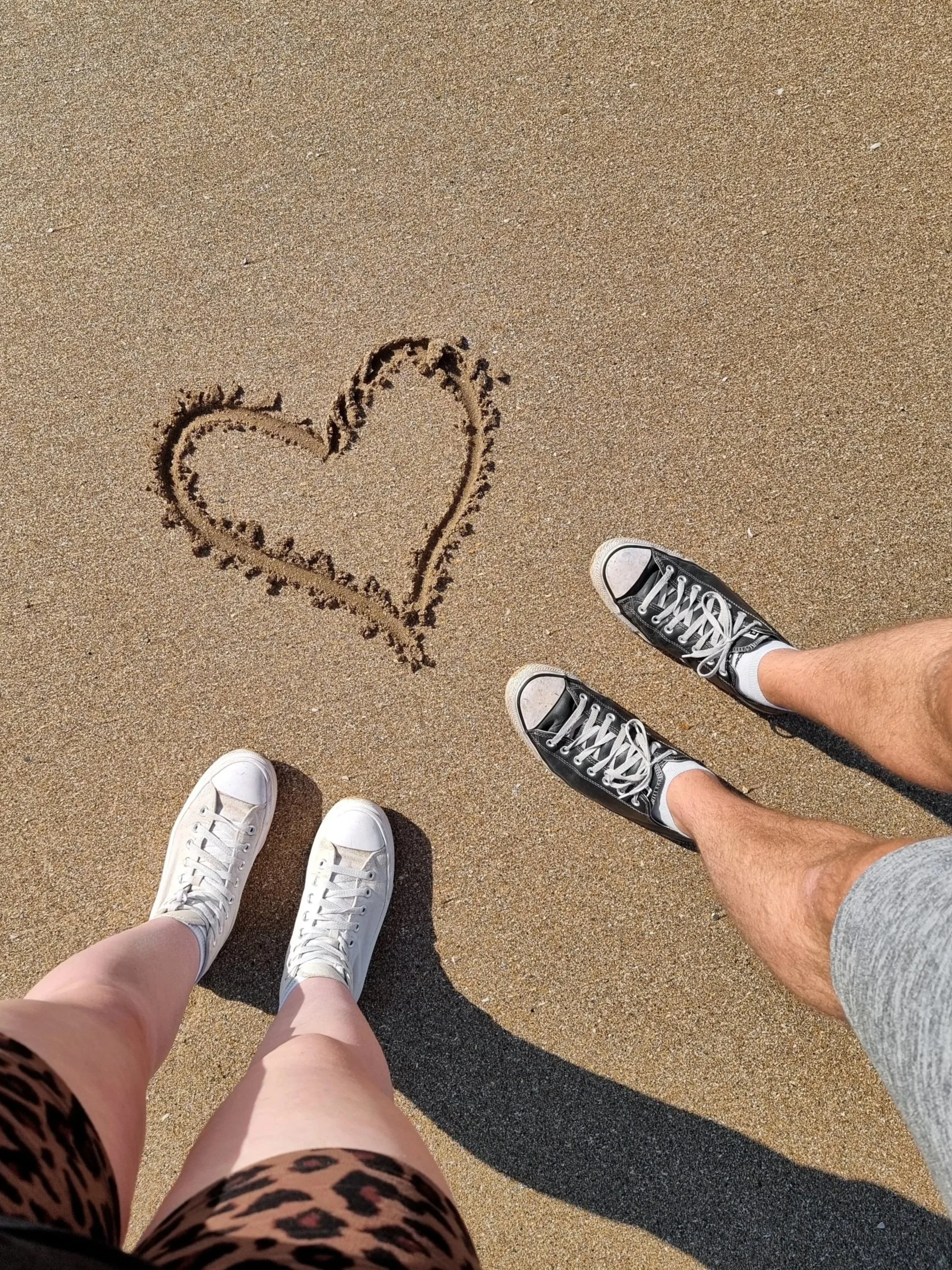 Two people standing on a sandy beach, one wearing white sneakers and pink shorts, the other wearing black sneakers and gray shorts, with a heart drawn in the sand.