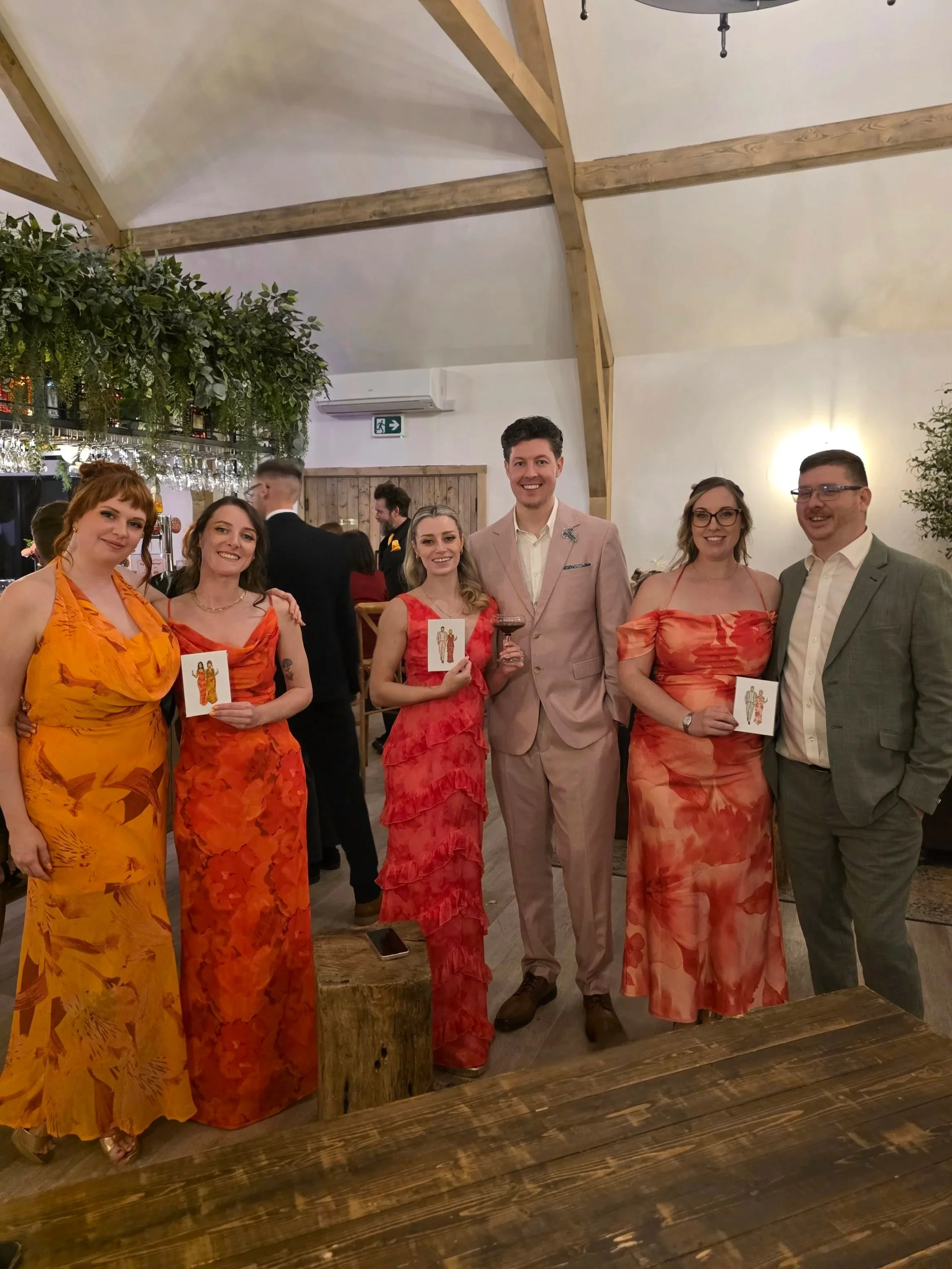 Group of six people posing at a social event, wearing colorful dresses and suits, holding small cards, standing in a warmly lit room with wooden beams on the ceiling and greenery decorations.