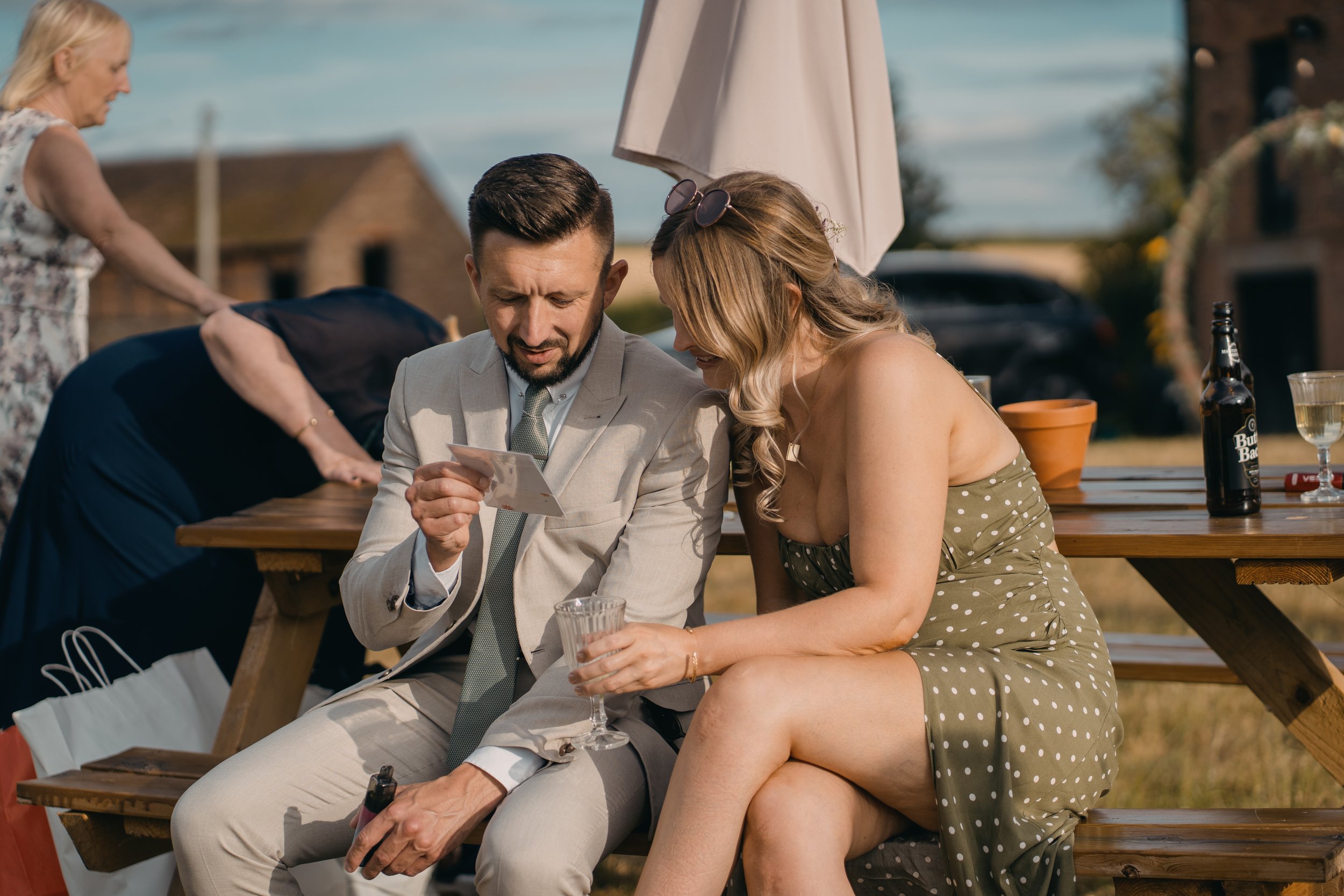 A man in a light grey suit and a woman in a green polka dot dress sitting closely at an outdoor gathering, looking at a phone together. There is a woman in the background setting up a table with drinks and a white umbrella overhead.