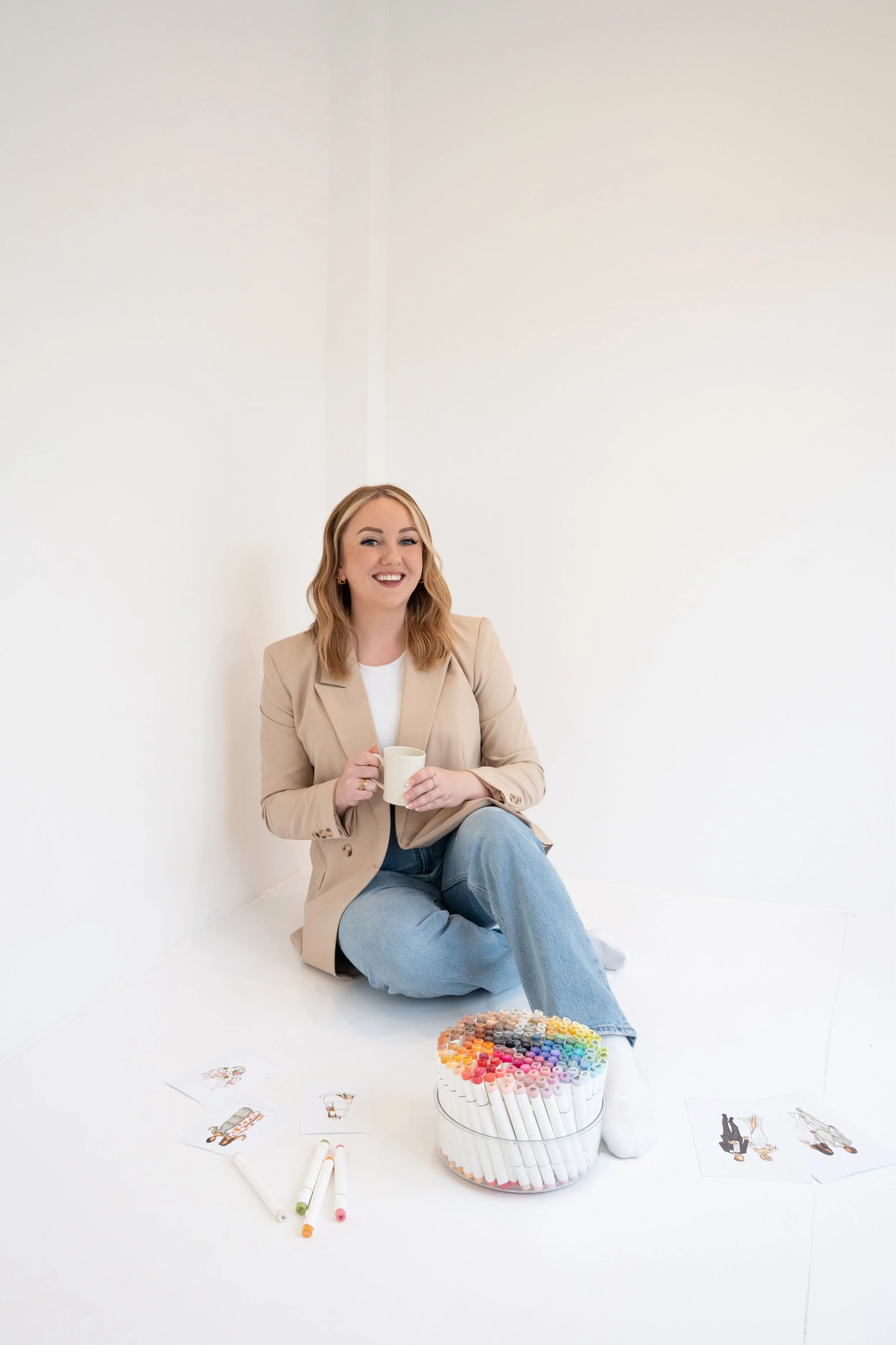 A young woman with blonde hair sitting on the floor against a white wall, smiling and holding a white mug. In front of her on the floor are colored markers, illustrated drawings, and a large container of colorful markers or pens arranged in a circular pattern.