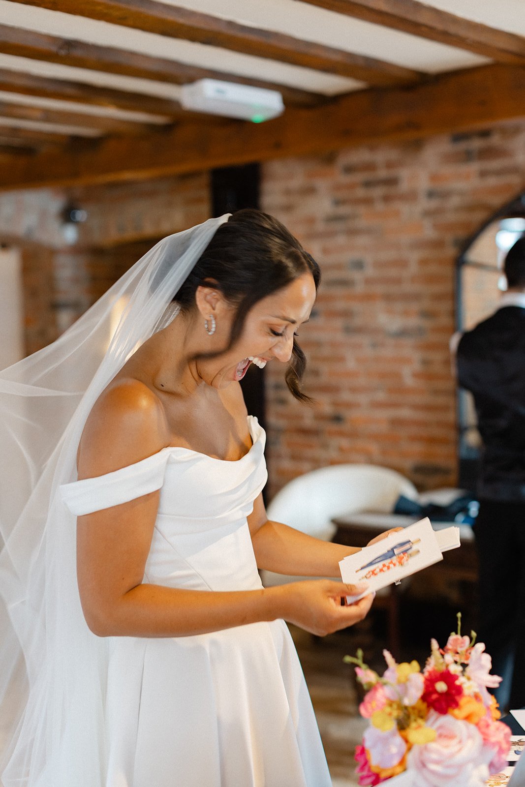 A bride in a white wedding dress and veil is smiling and reading a card at her wedding reception, with a cake with pink flowers in front of her and a man in a tuxedo in the background.