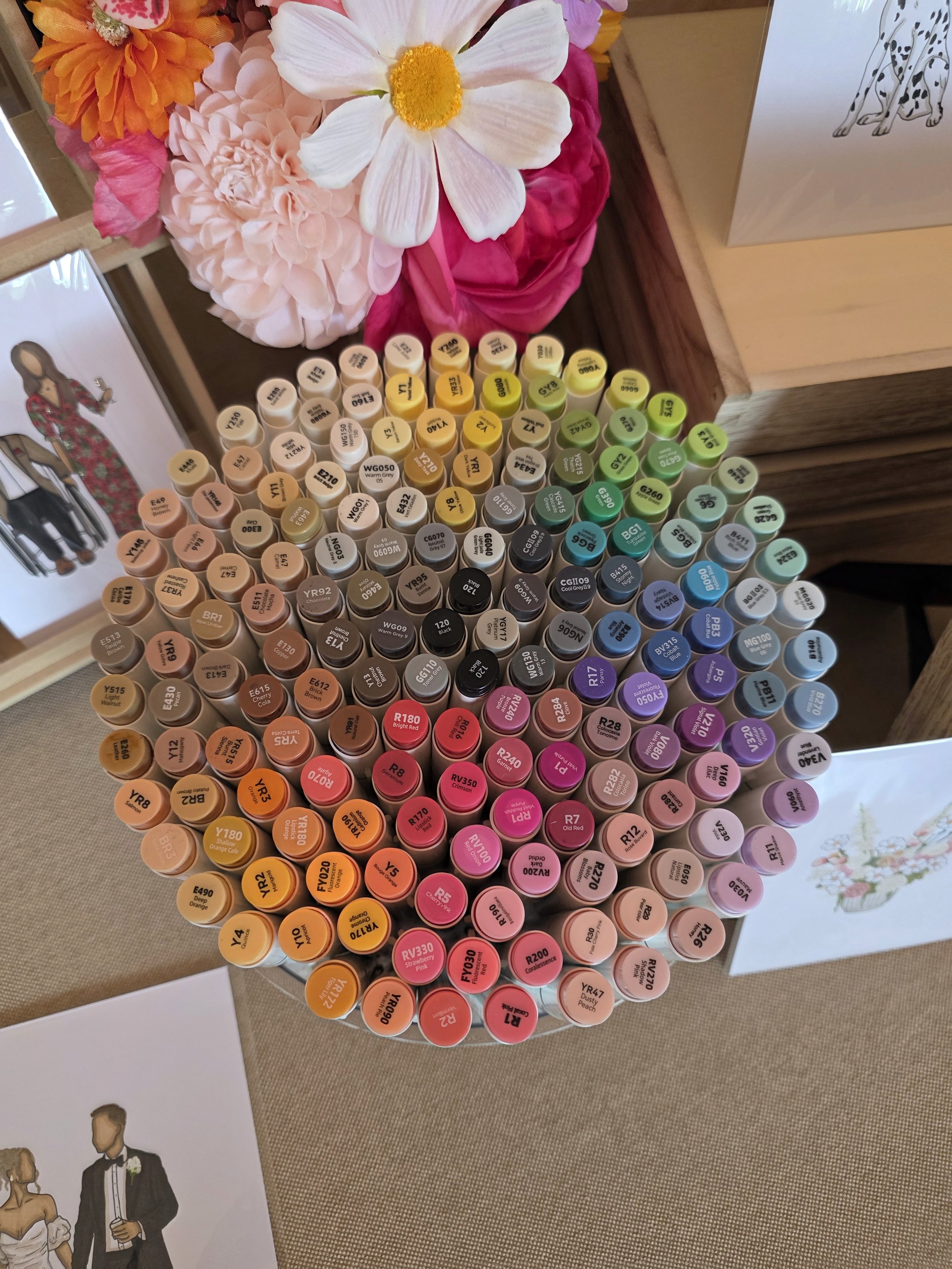 A collection of colorful paint marker pens arranged in rows with their color codes visible on the caps. There are shades of peach, pink, yellow, green, blue, purple, black, and brown. In the background, there are decorative floral arrangements and a drawing of a woman in a dress and a man in a tuxedo.
