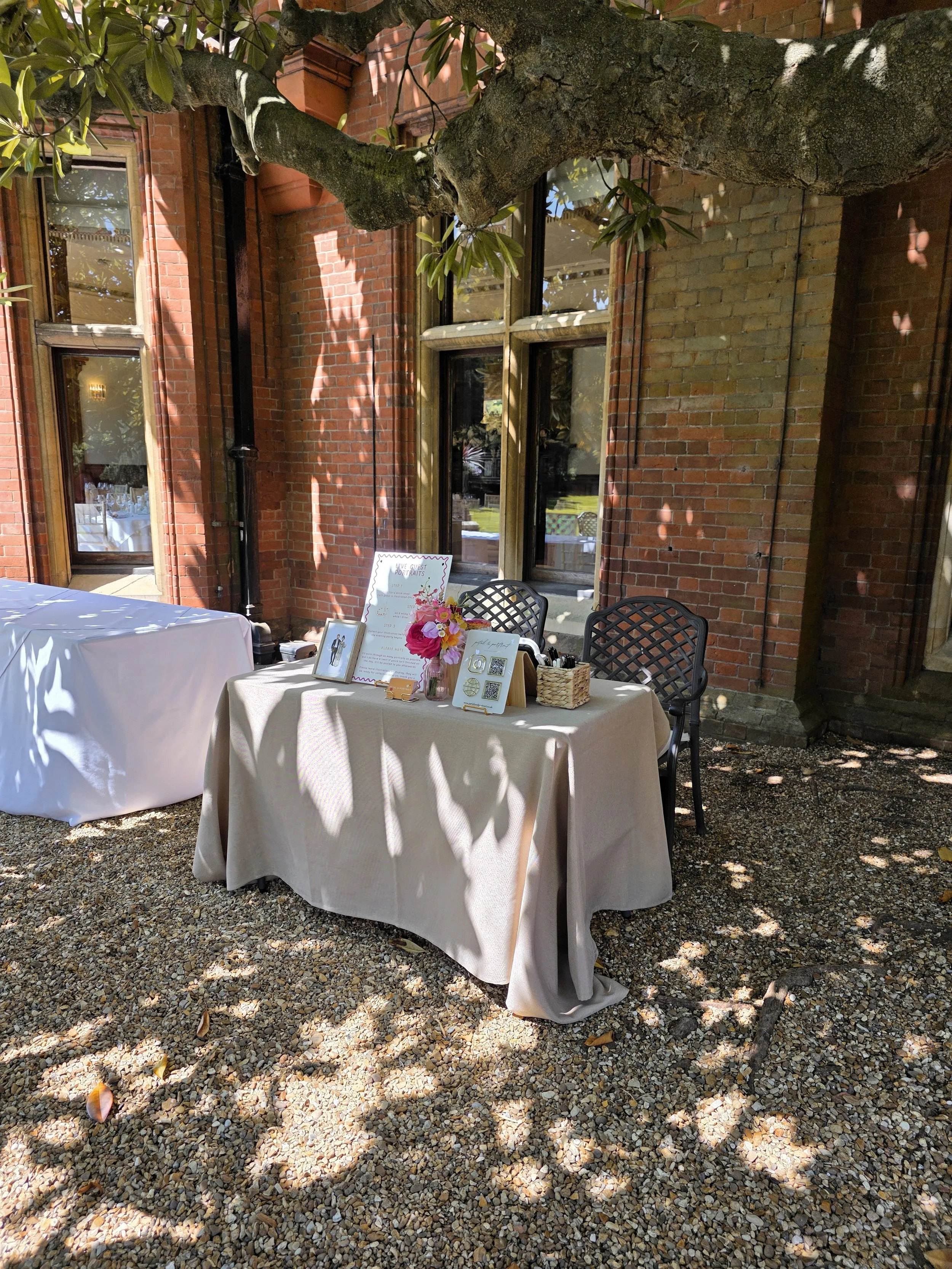 Outdoor party setup with two tables covered in beige and white tablecloths, decorated with flowers, picture frames, and signs next to a brick building and a tree with branches overhead.