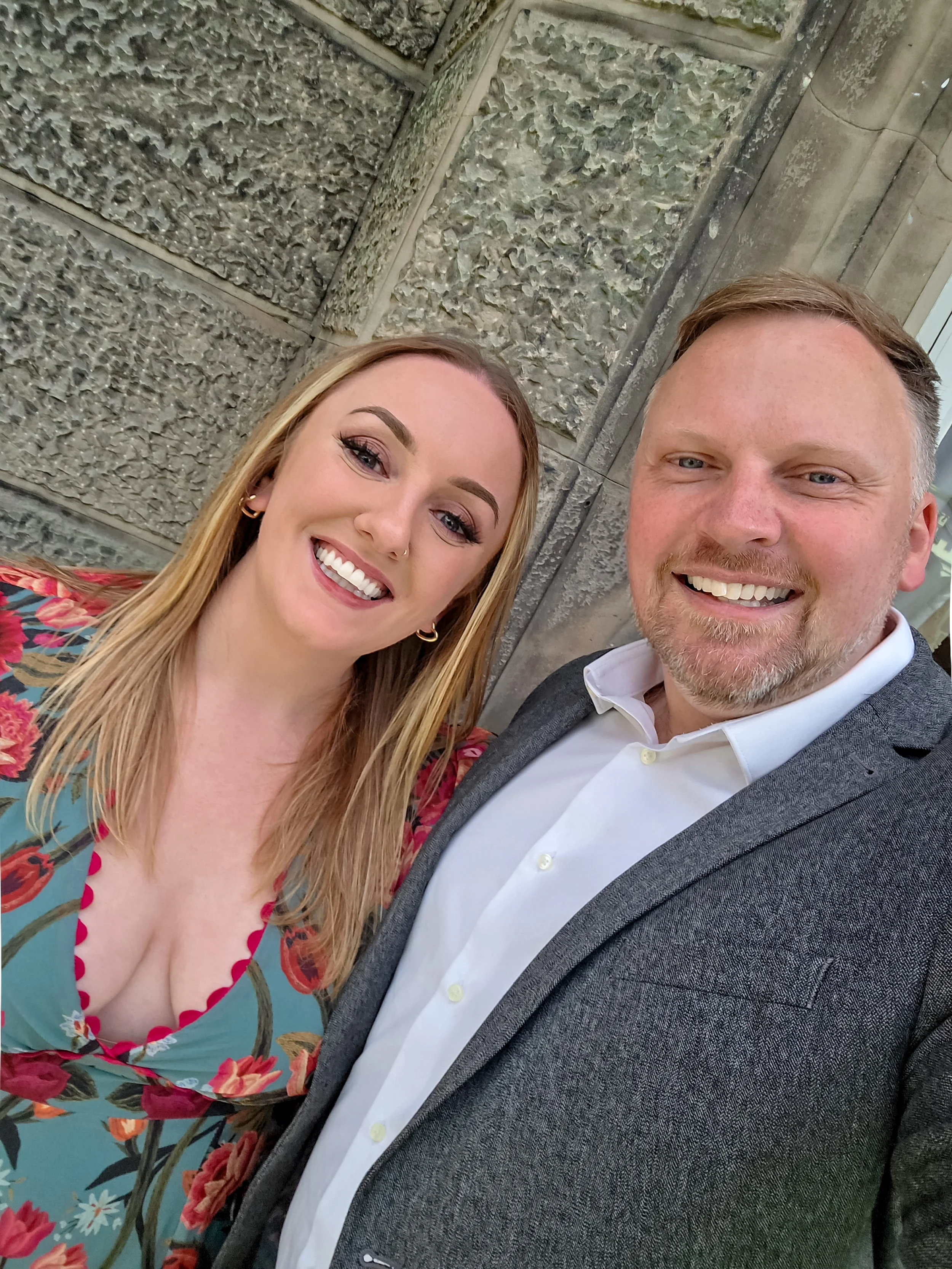 A smiling woman with long blonde hair and a man with short light brown hair and a beard, both dressed in formal attire, standing closely together in front of a stone wall, taking a selfie.