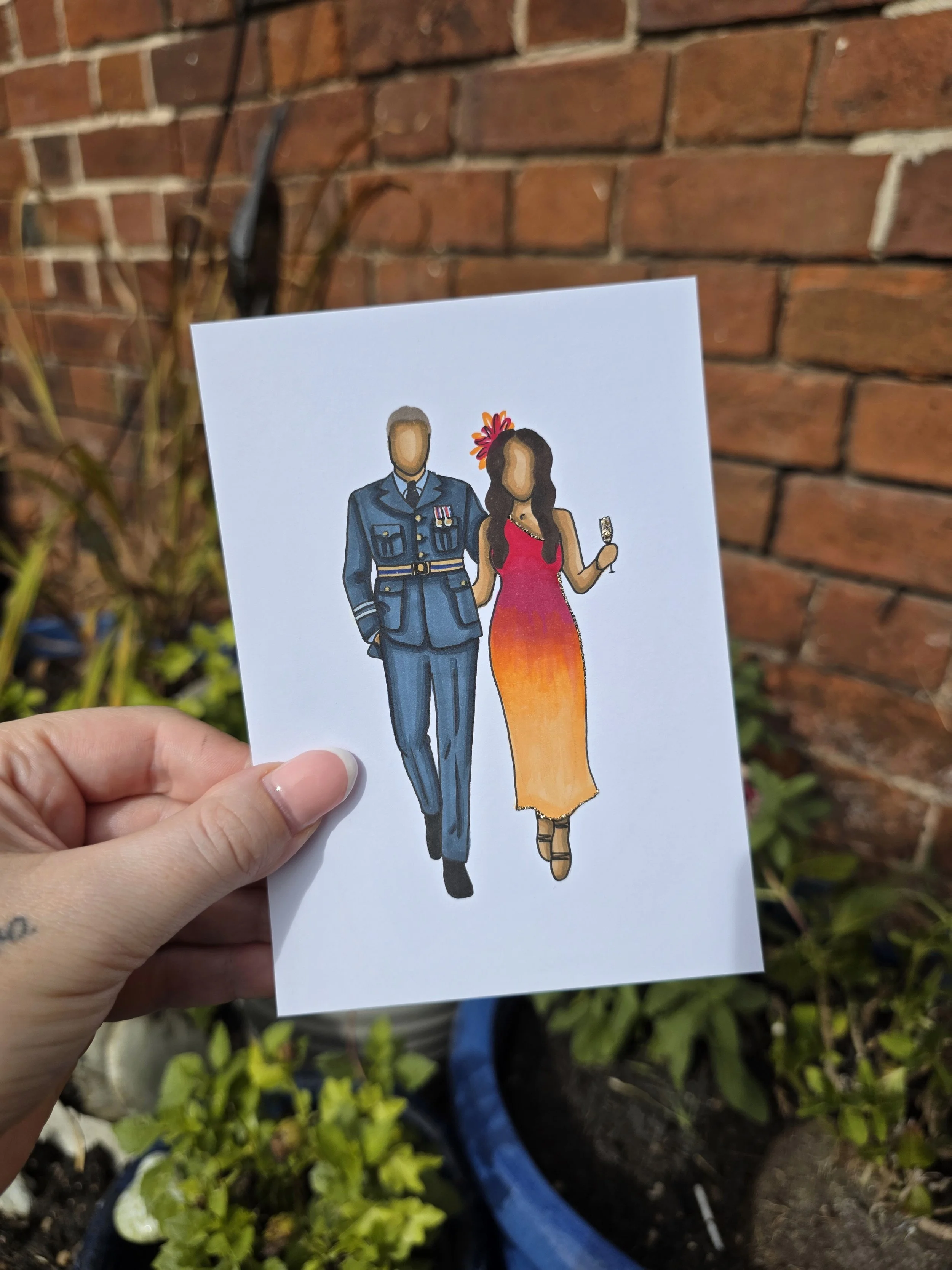 A hand holding a drawing of a man in a blue military uniform and a woman in a colorful dress with a flower in her hair, against a brick wall background.