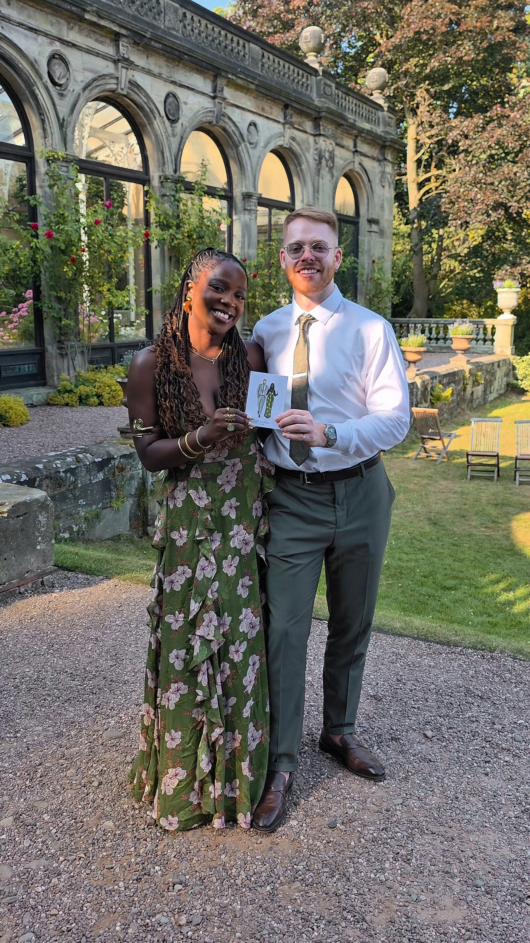 A smiling Black woman with long braided hair and gold jewelry standing next to a smiling White man with glasses, dressed in a white shirt and tie, in a garden with a glass greenhouse and trees in the background, holding a small photo or card.
