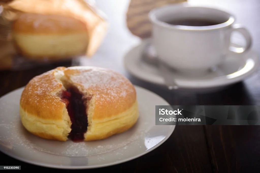 A fluffy cake with a cut revealing a berry filling, dusted with powdered sugar, on a white plate next to a cup of black coffee on a saucer.