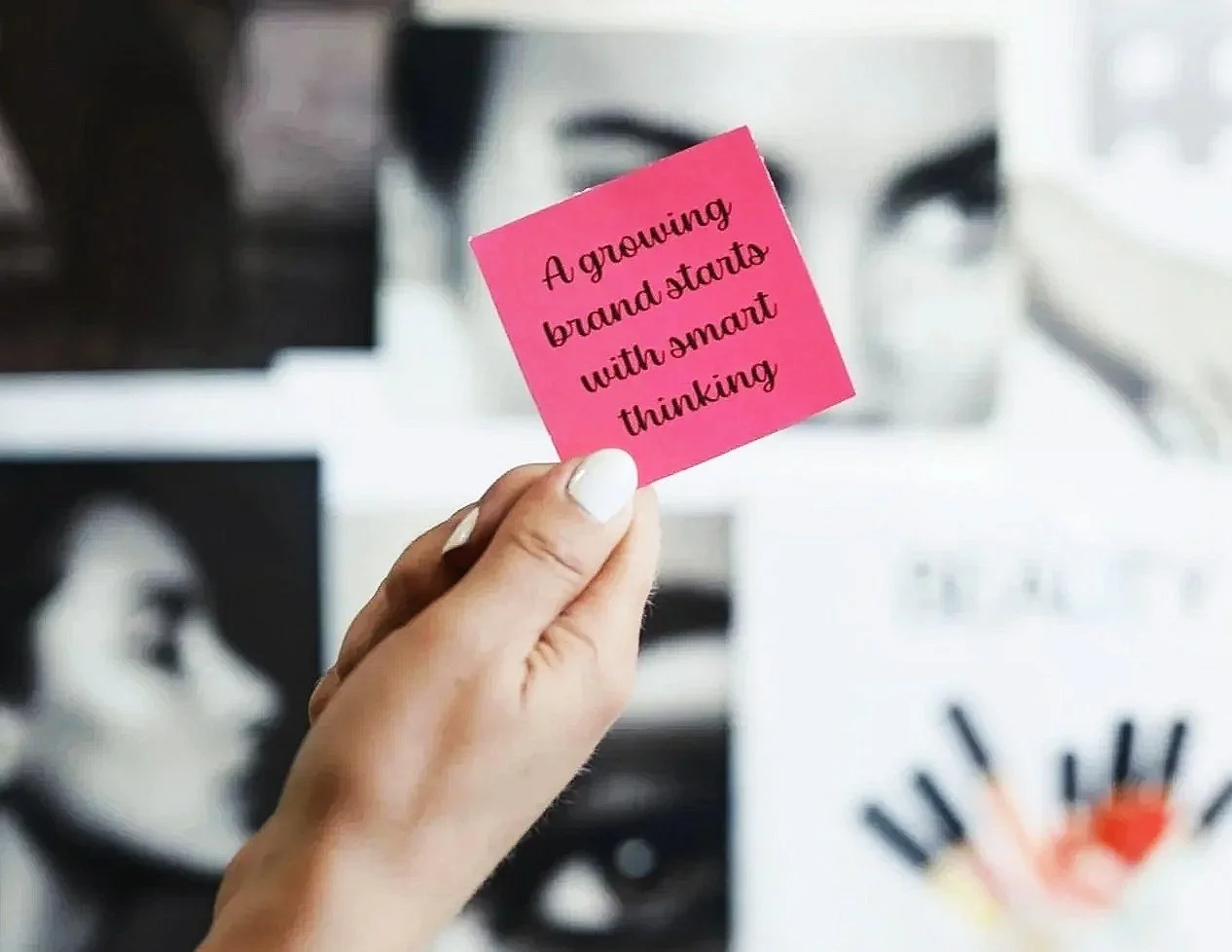 A hand holding a pink sticky note that reads 'A growing brand starts with smart thinking'. The background includes blurred images of colorful pens and a black-and-white artwork.